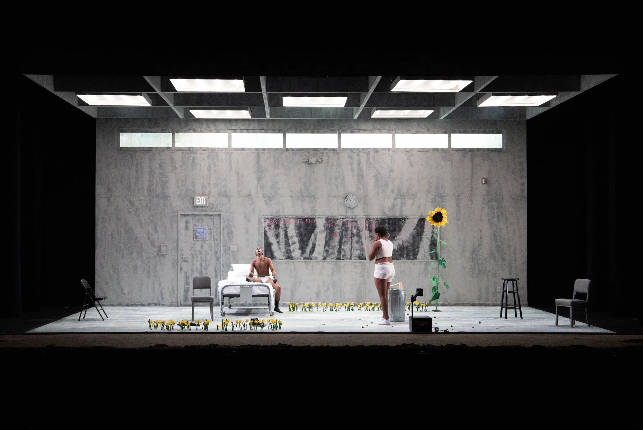Theatrical stage set of a minimalist room with concrete walls, a bed, chairs, and a large sunflower. Two people are on stage in casual attire, with flowers on the floor, creating an abstract scene.