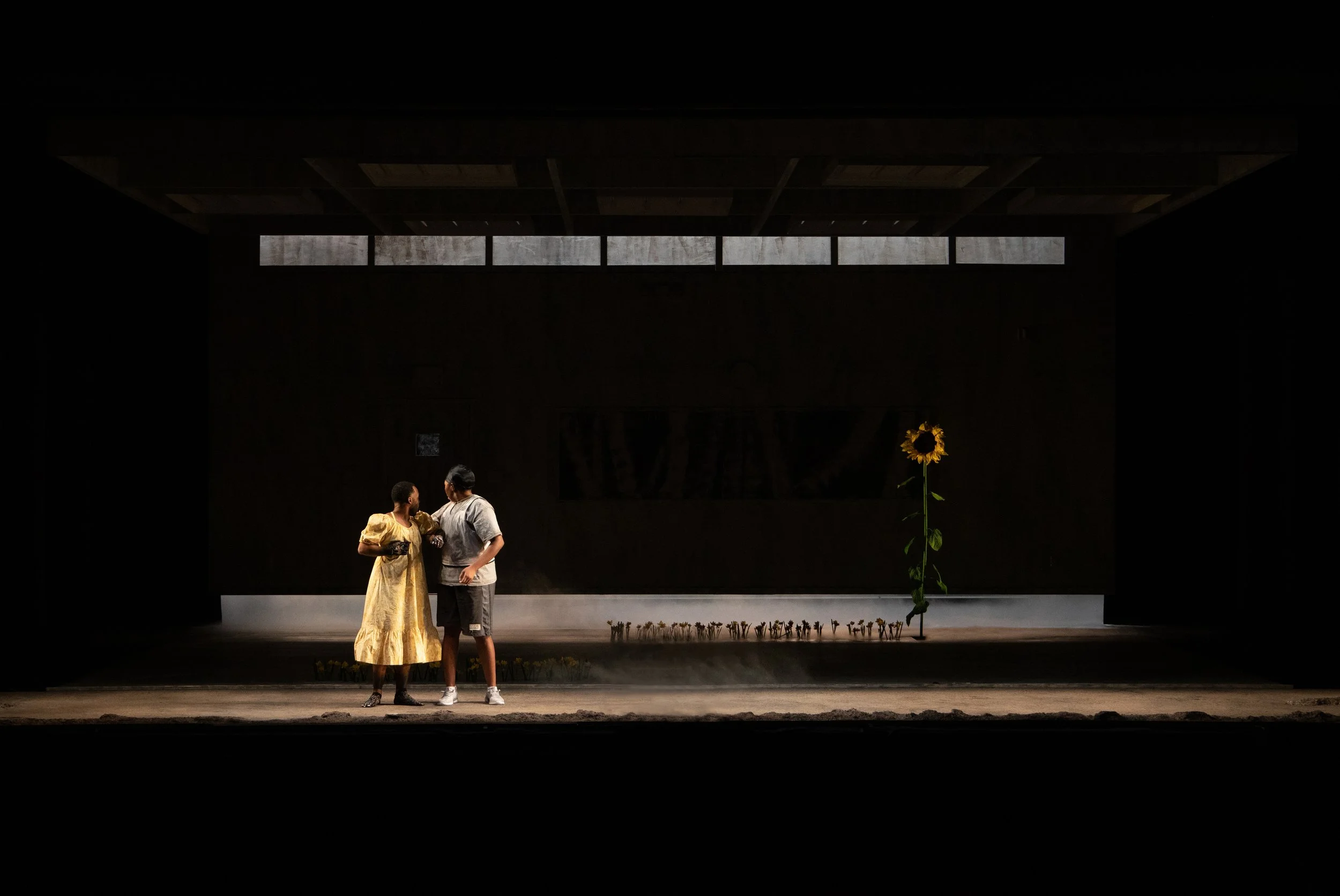 Two people standing on a dimly lit stage with a large sunflower in the background.