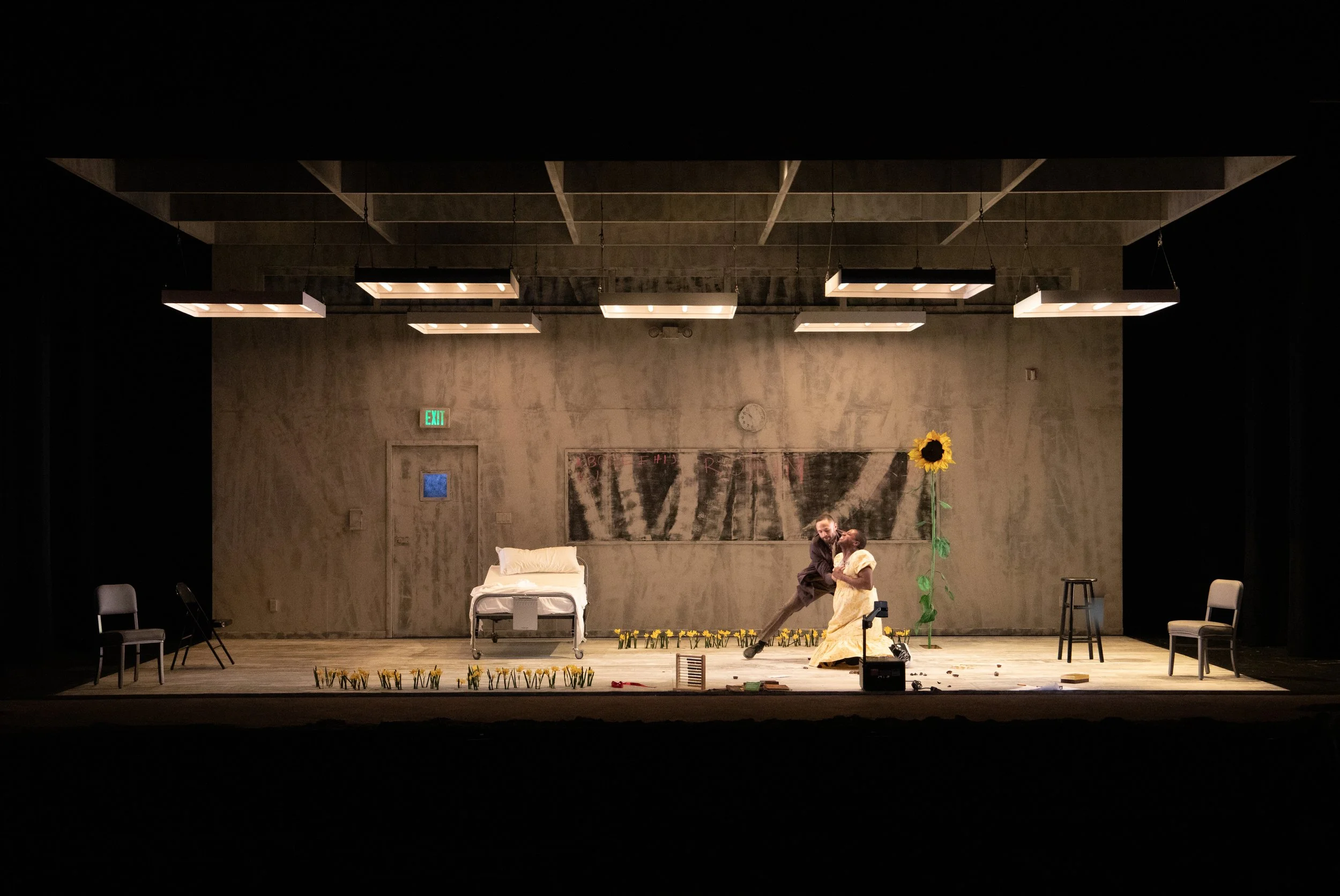 Theater stage set with a hospital bed, chairs, large sunflower, two people in dramatic pose, and dim overhead lighting.