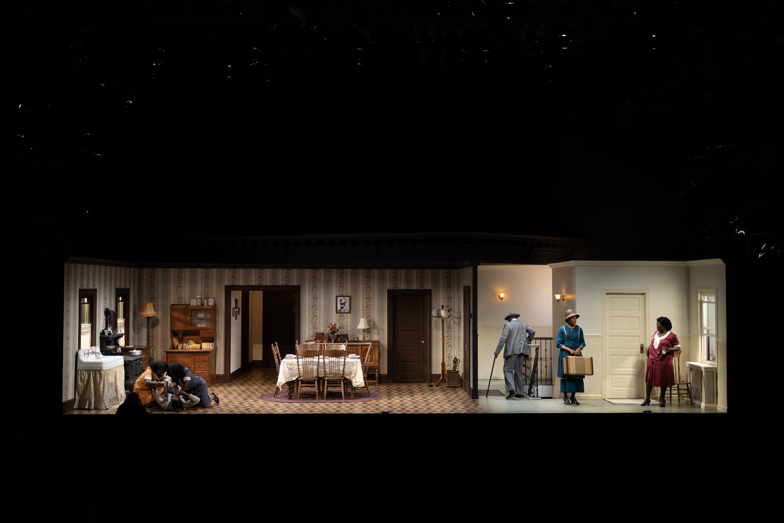 A theatrical stage set depicting a mid-20th-century living room and hallway. On the left, two people are sitting on the floor of a cozy, patterned wallpaper room with a dining table. On the right, a hallway shows two people standing near a door. One 