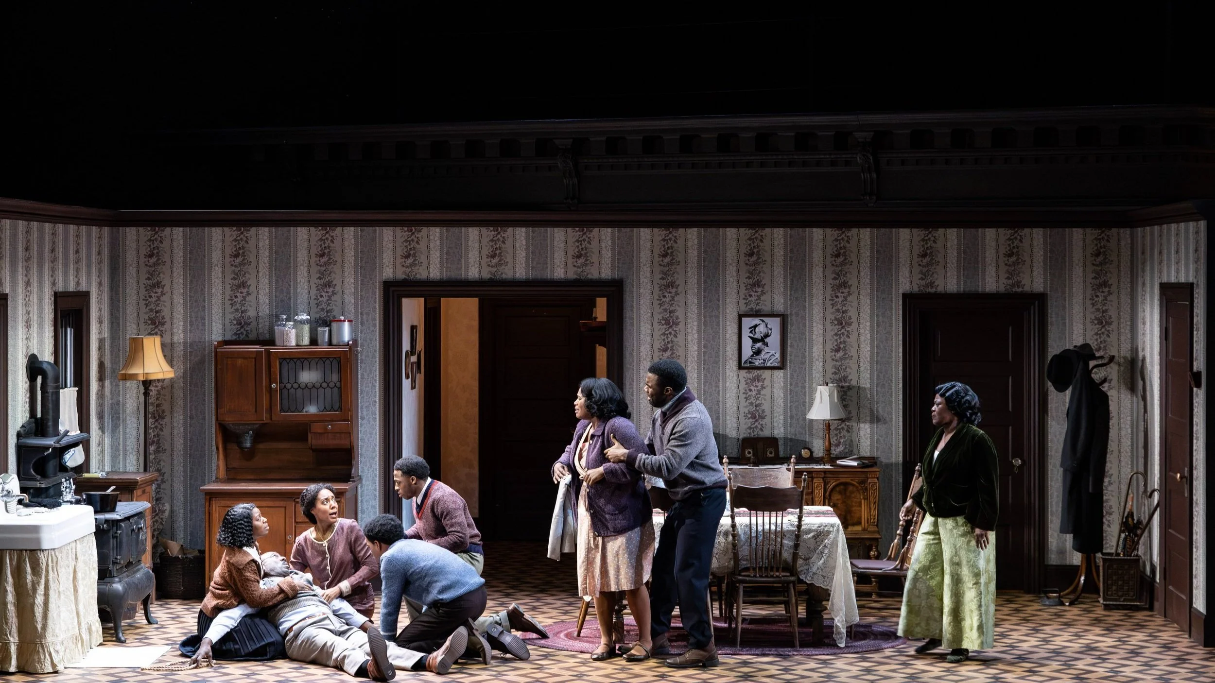 Stage scene depicting a vintage living room with actors portraying a dramatic moment. One person is lying on the floor surrounded by others, who display concern. The set features period furniture, a stove, and floral wallpaper.