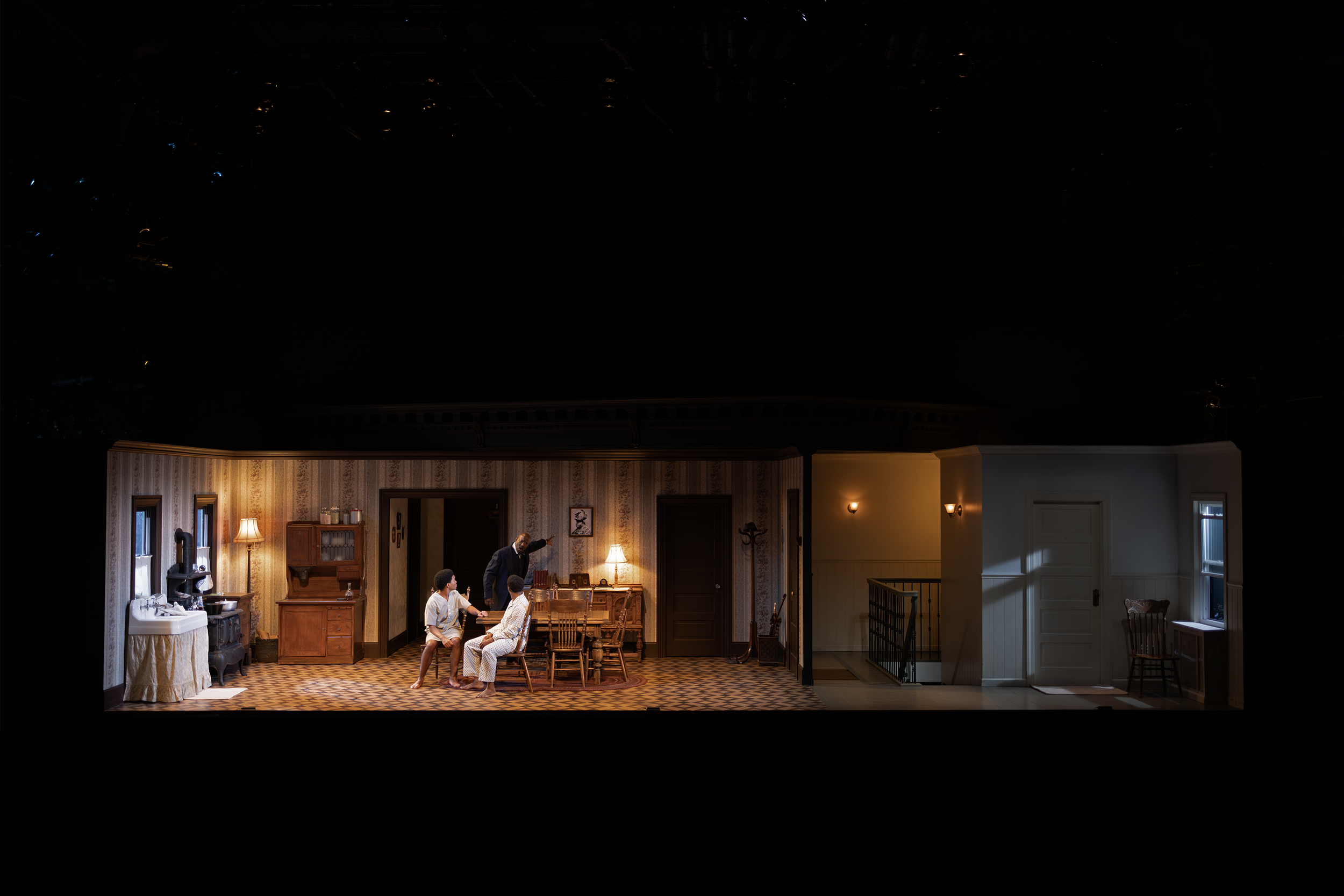A dimly lit theater stage set depicting an interior scene with a vintage-style living room and kitchen. Three people are interacting at a dining table. The scene includes period furniture, wallpaper, and decor, with a side area that appears to lead t