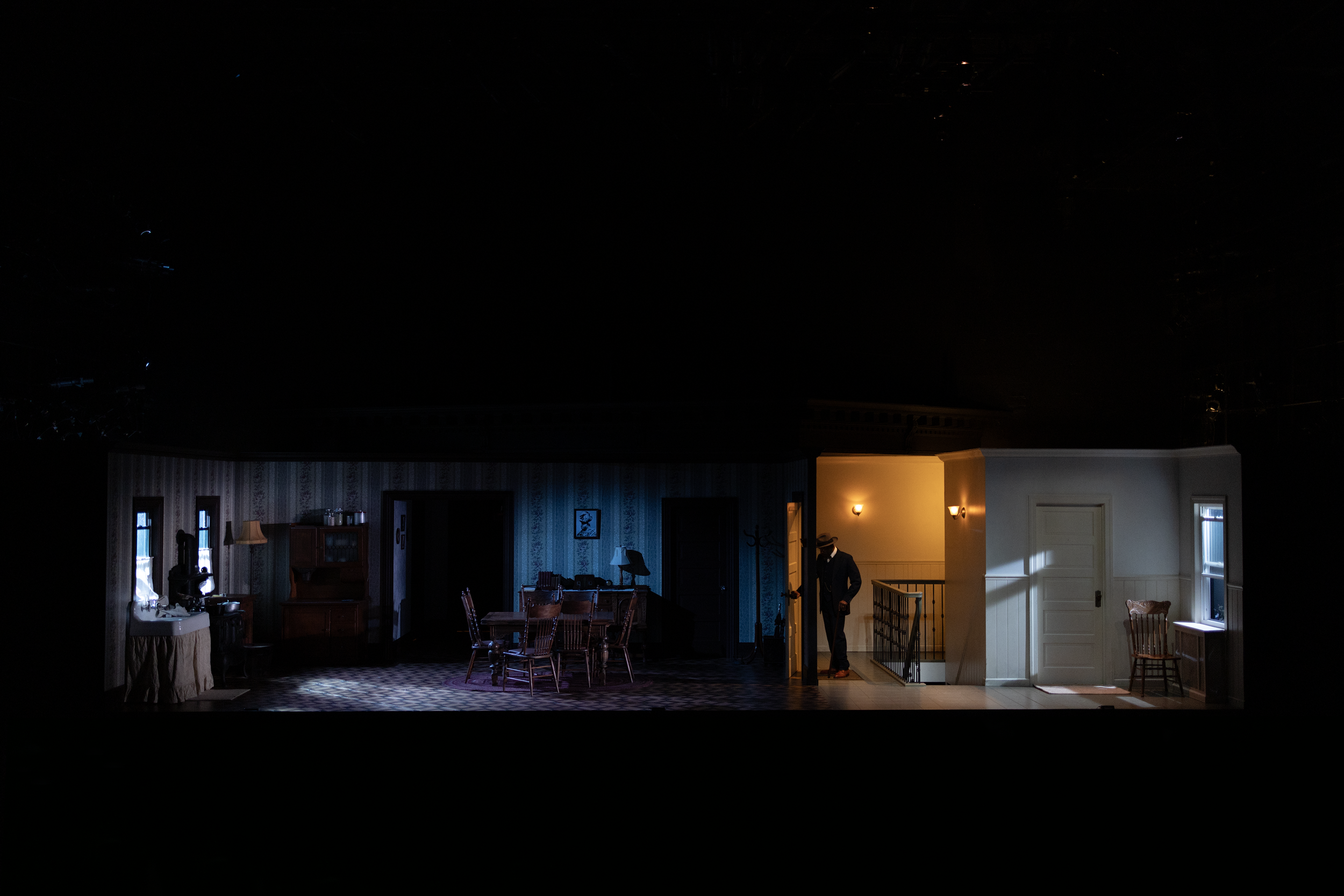 Dimly lit theater stage set depicting an early 20th-century home with vintage furniture, including chairs and a table, and a character in a suit standing near a door.