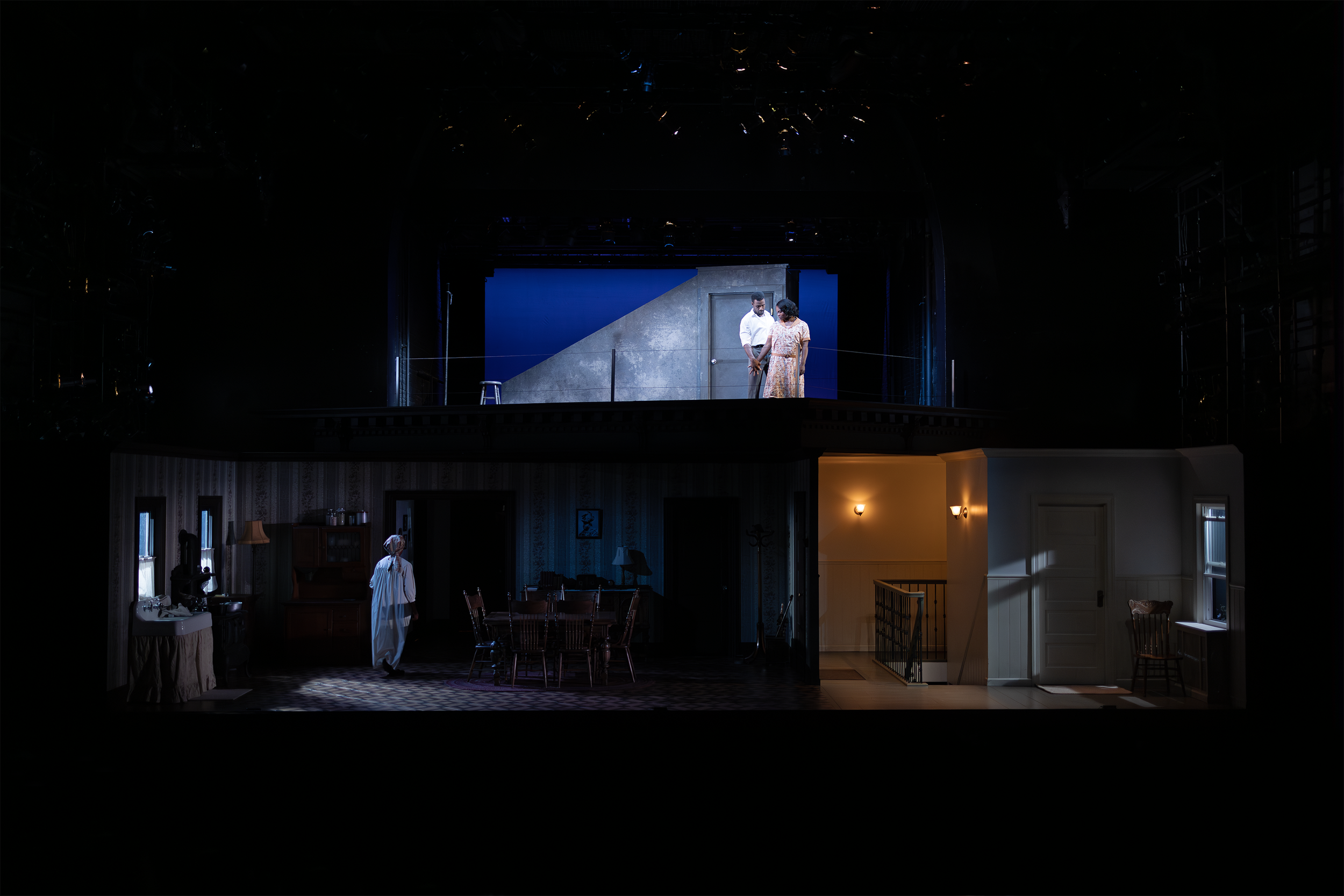 Stage production scene showing a two-level set with actors. The lower level depicts an interior room with furniture, while the upper level features two people standing outdoors against a blue sky backdrop. The setting includes moody lighting, suggest