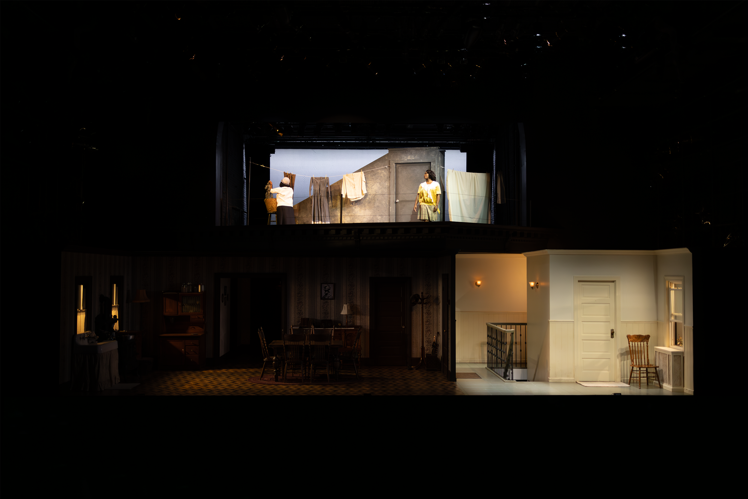 Theater stage set depicting a two-level scene; top level shows two actors hanging laundry on a rooftop, with clotheslines and a gray background. The lower level shows an interior home setting with a dining room and another room with a staircase and c