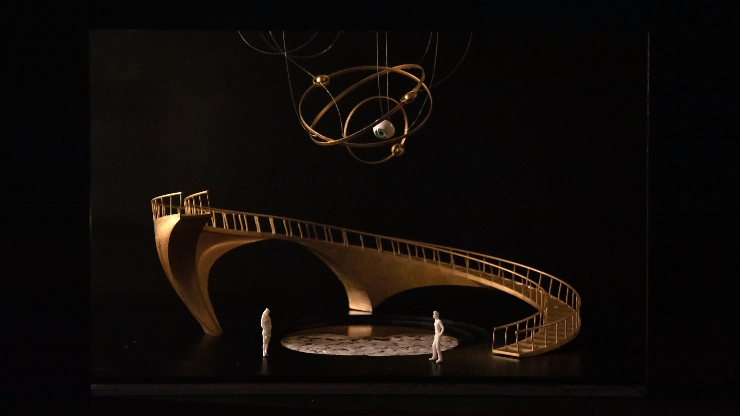 Miniature art installation featuring a golden spiral staircase and an abstract mobile with rings and a spherical eye, set against a dark background.