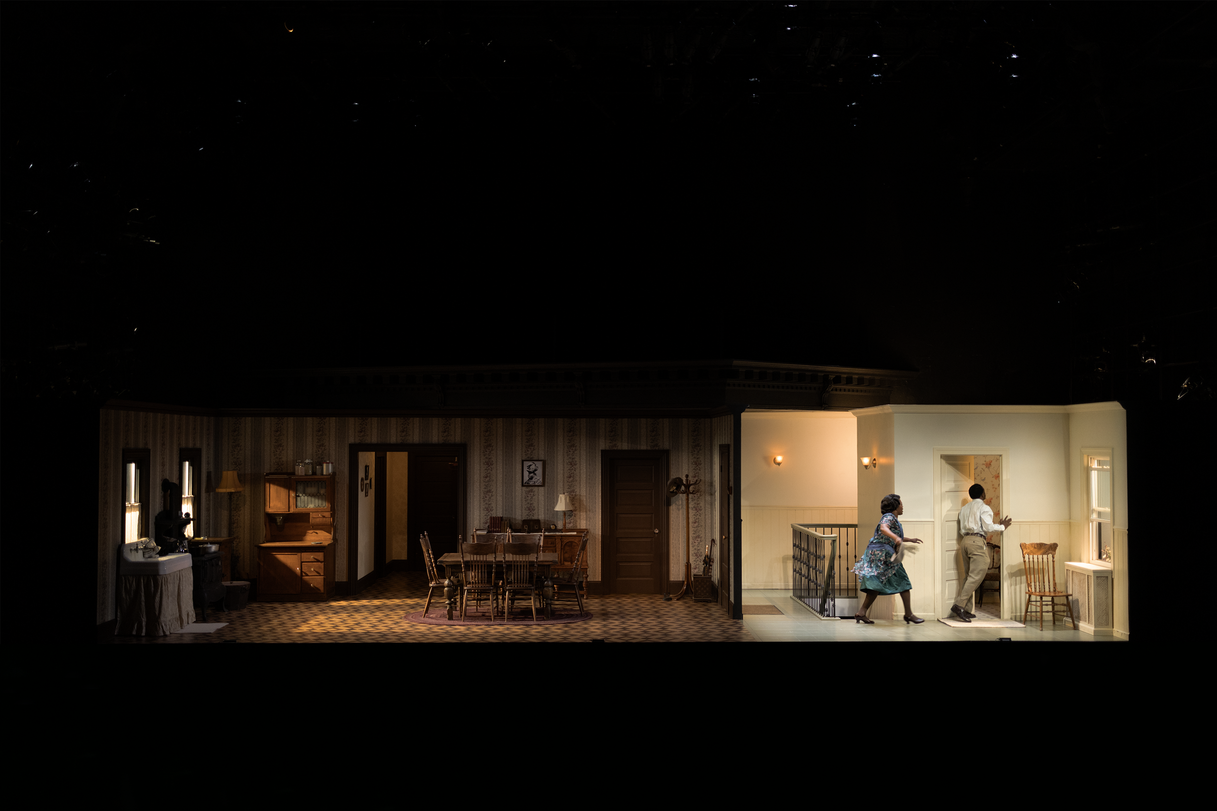 Stage set with two actors in a domestic interior, featuring a dining area, kitchen, and hallway, with dim lighting and period-appropriate furniture.
