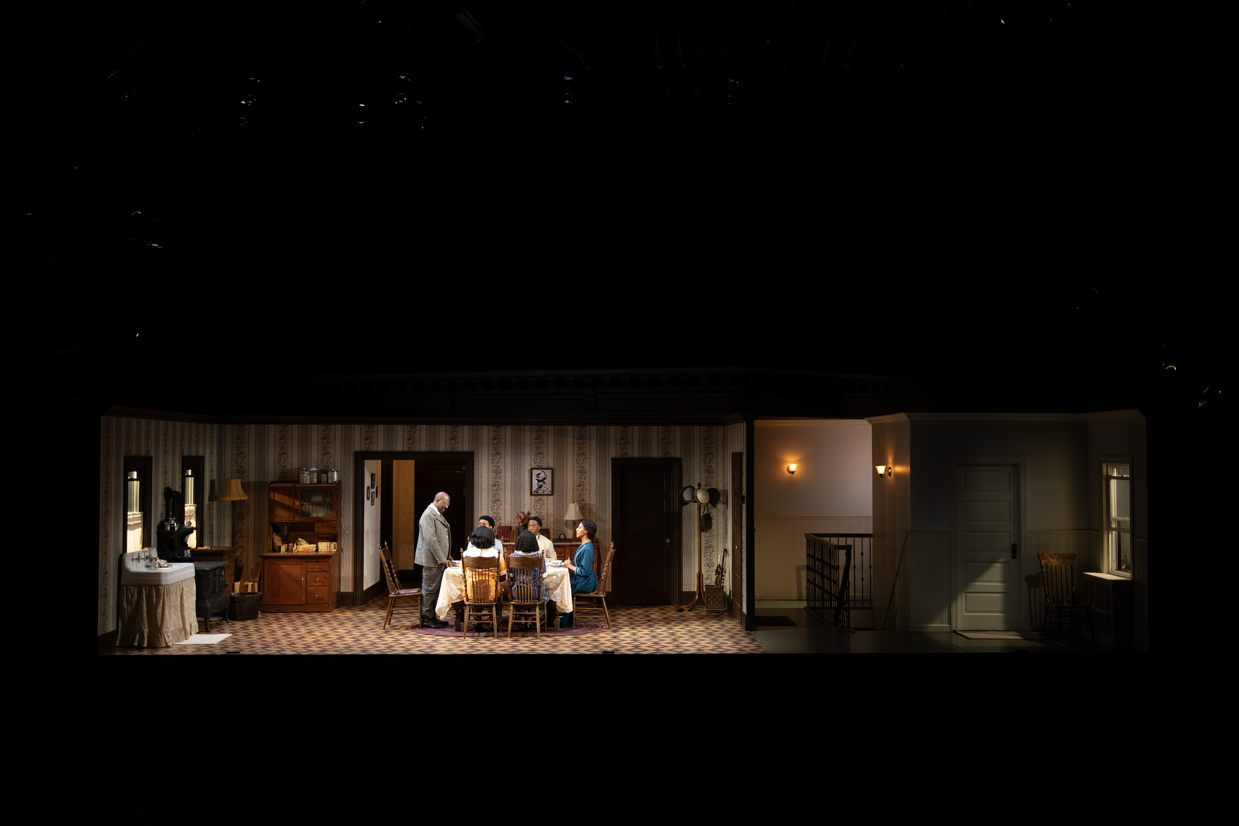 A stage set depicting an interior of a house with five people seated around a dining table. The room has period-style decor and furniture. A man is standing at the head of the table, engaging with the seated individuals. The scene is dimly lit, creat