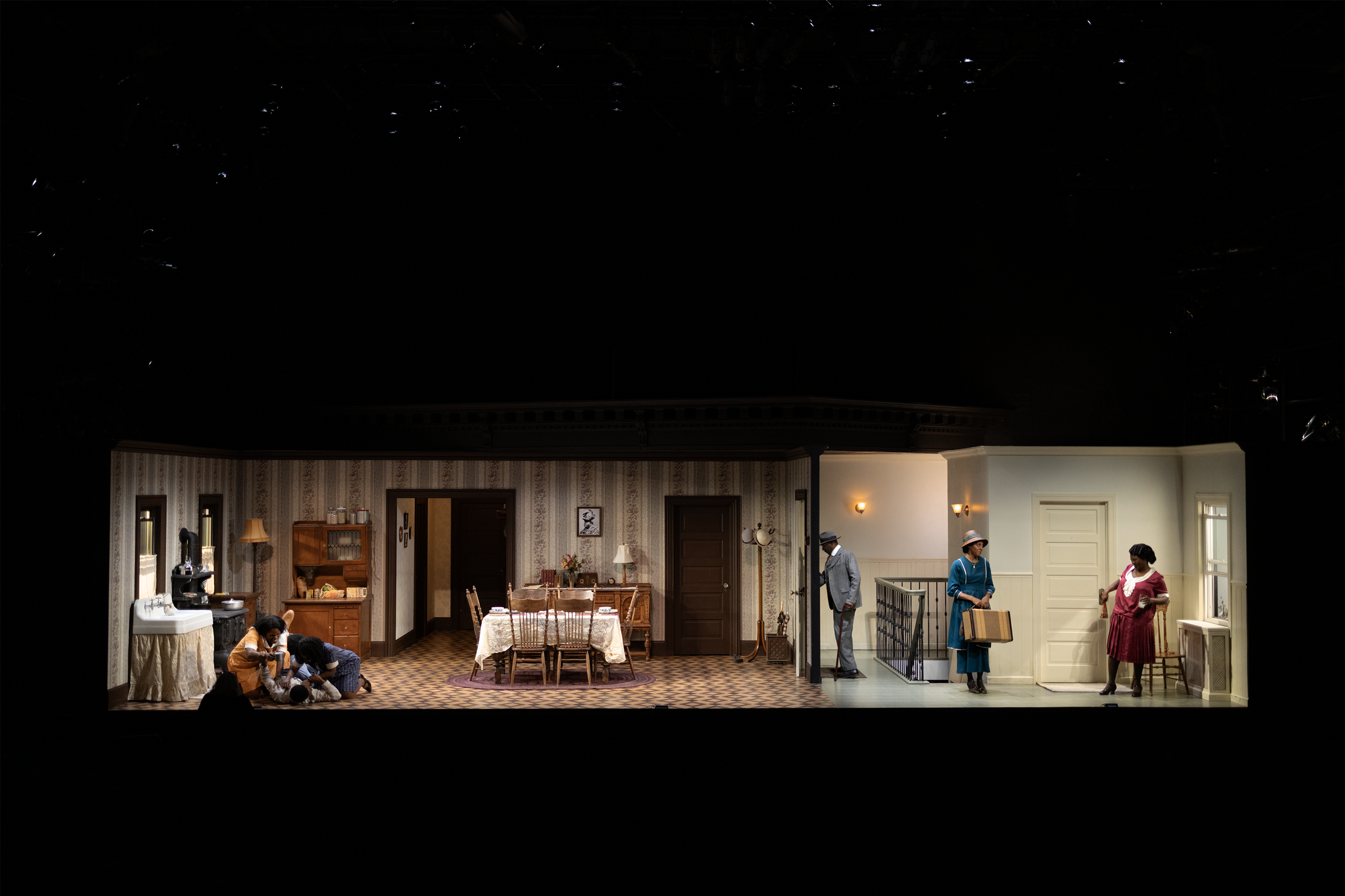 Stage set of a theater scene depicting a domestic interior with people. The scene is split into different rooms, showing characters engaged in various activities. On the left, a kitchen with a table and chairs, and two people kneeling over a dog. On 