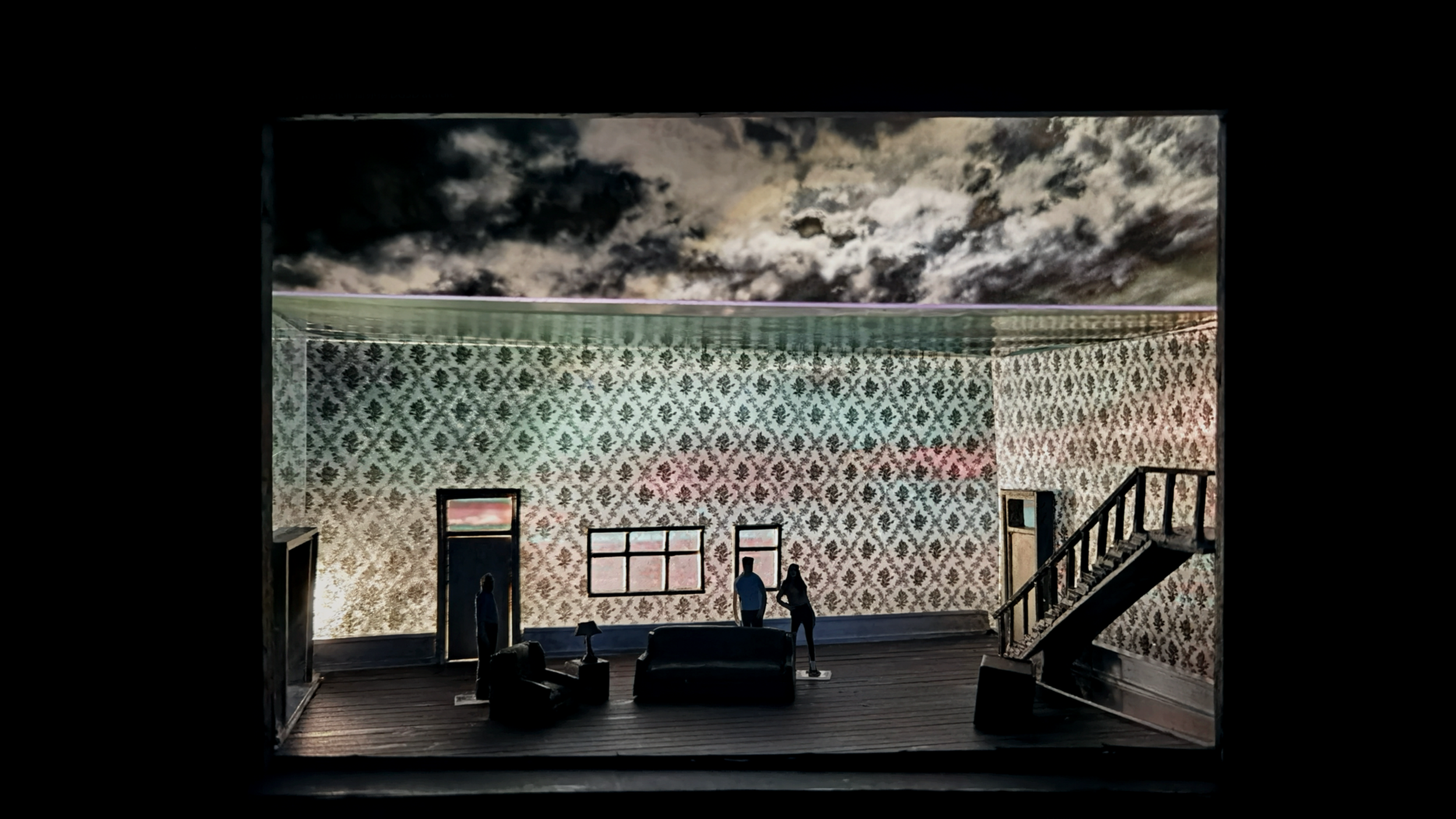 Miniature diorama of a vintage living room with patterned wallpaper, a couch, chairs, a lamp, and stairs; silhouetted figures by windows with a stormy sky background.