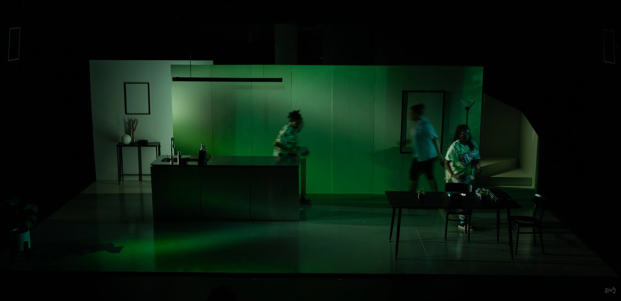 Dimly lit stage scene with green lighting, featuring several figures in motion around a minimalist kitchen and dining room setup, including a central table and chairs, a kitchen island with a sink, and wall decor.