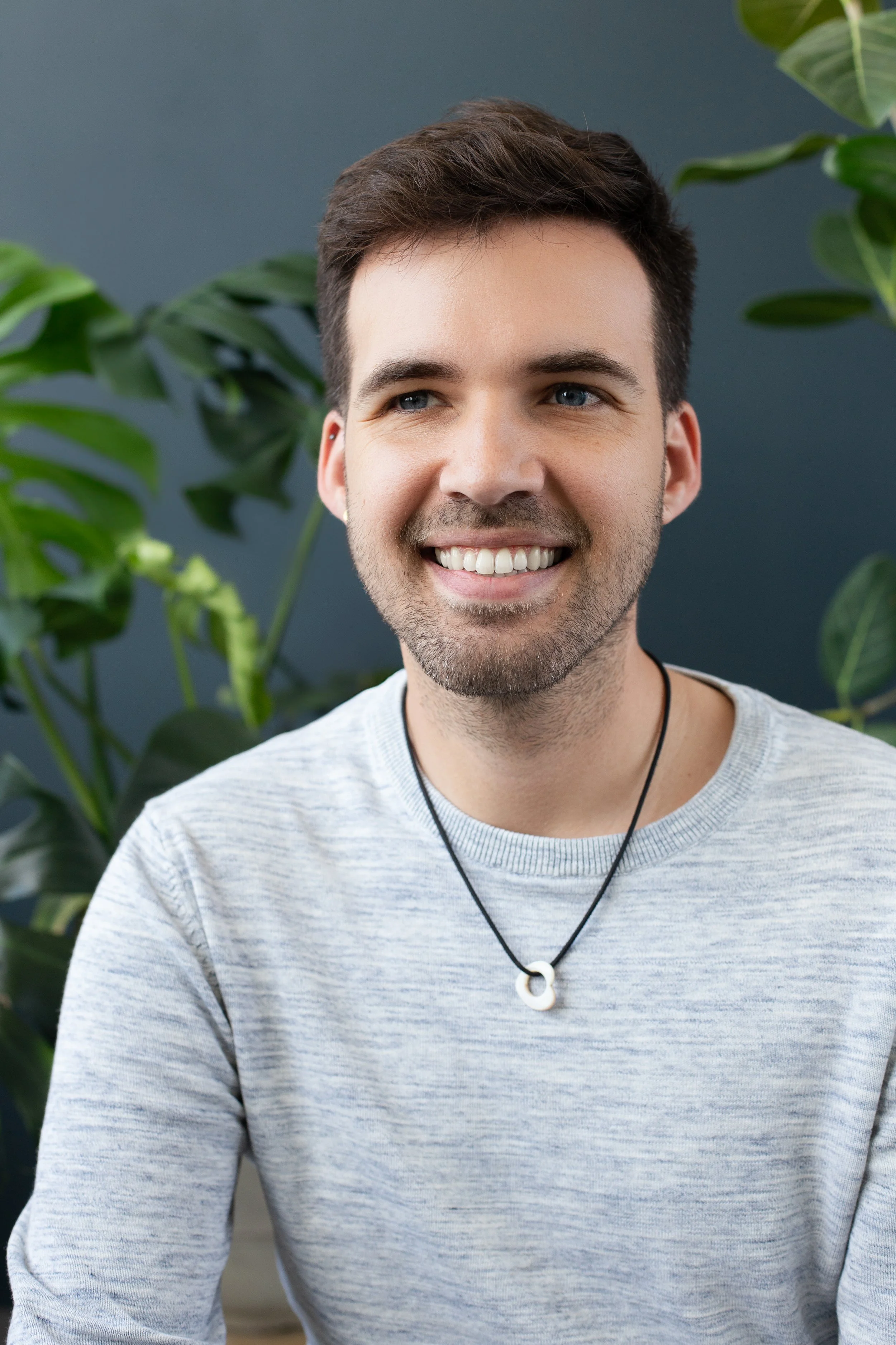 A smiling young man with short dark hair, a beard, and blue eyes, wearing a light gray shirt and a black necklace with a white circular pendant, seated in front of green indoor plants on a dark blue wall.