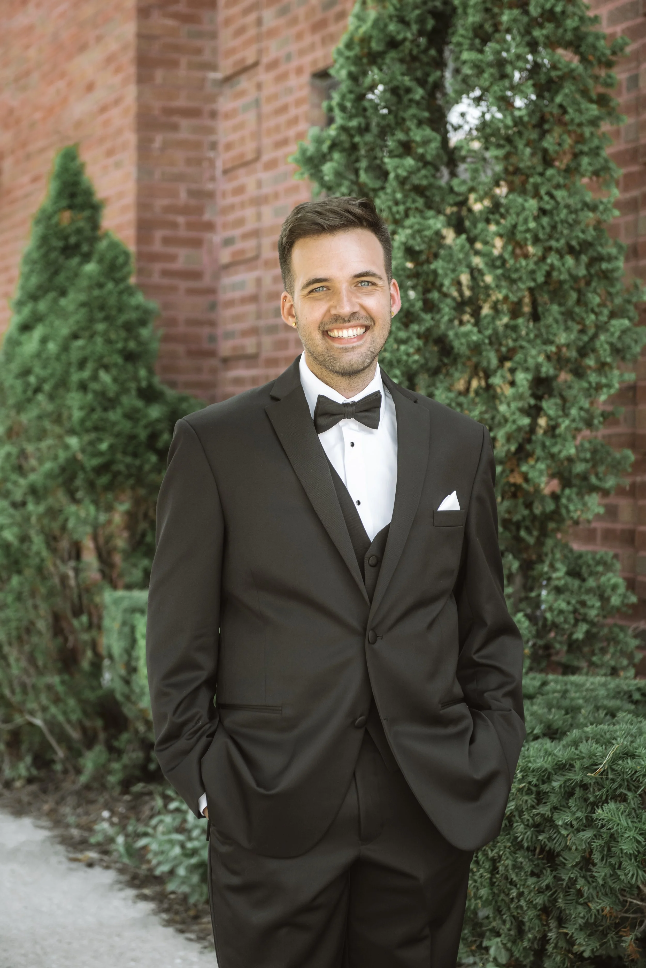 A smiling man dressed in a black tuxedo with a bow tie, standing outdoors with green bushes and trees behind him.