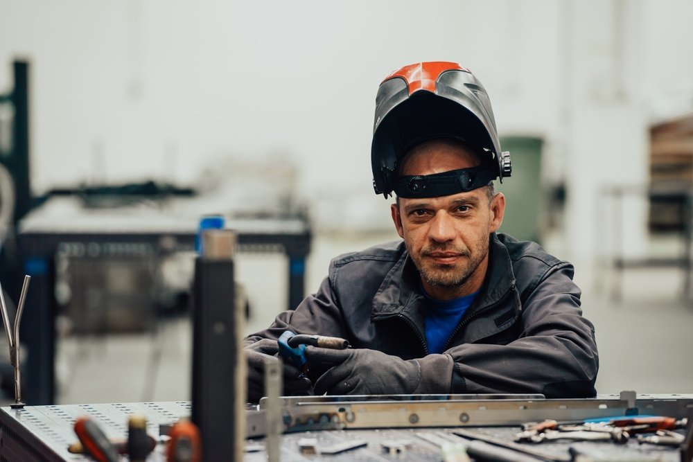 A man wearing a welding helmet lifted on his head, sitting at a workbench with tools in a workshop. He is upset with his working conditions and has decided to look for a new job. A friend of his told him about weldingrecruiter.com.