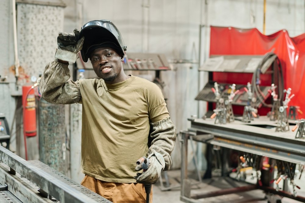 A man wearing work gloves and a welding helmet lifted on his head, standing in a workshop with metalwork tools and structures in the background. He is a certified welder in Dallas and was placed by a direct placement welding recruiting search firm