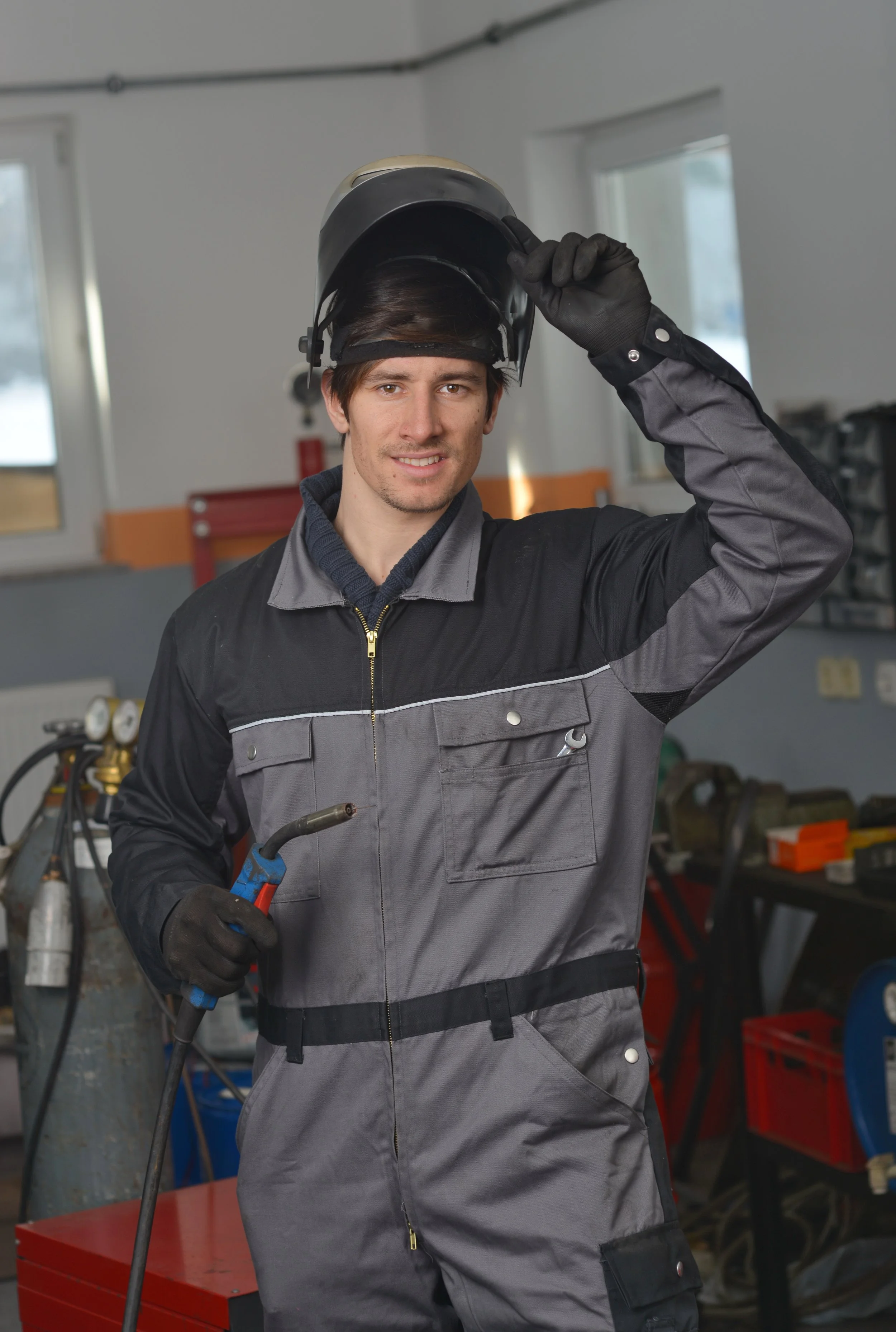 A young man in a gray and black work jumpsuit is smiling and adjusting his protective welding helmet while holding a welding tool in his gloved hand inside a workshop.