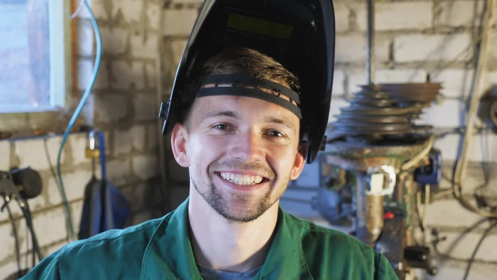 A young man wearing a welding helmet is smiling because he knows companies are having trouble finding certified welders, and he just found out his value on the market.