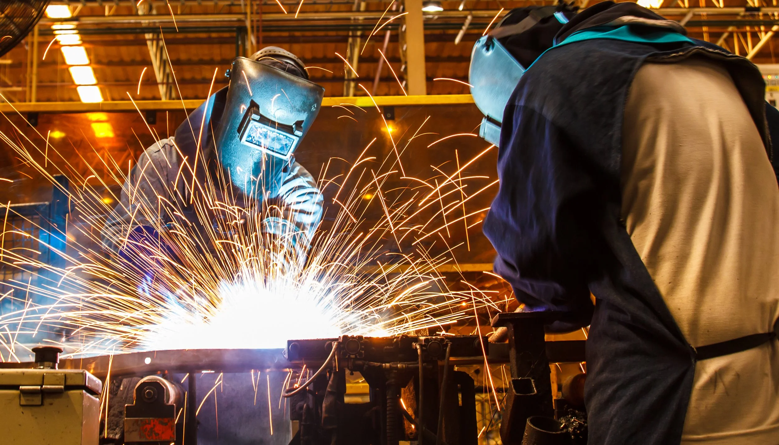 Two workers welding metal with sparks flying in a factory workshop. They have both recently interview with a direct hire welding recruiter.
