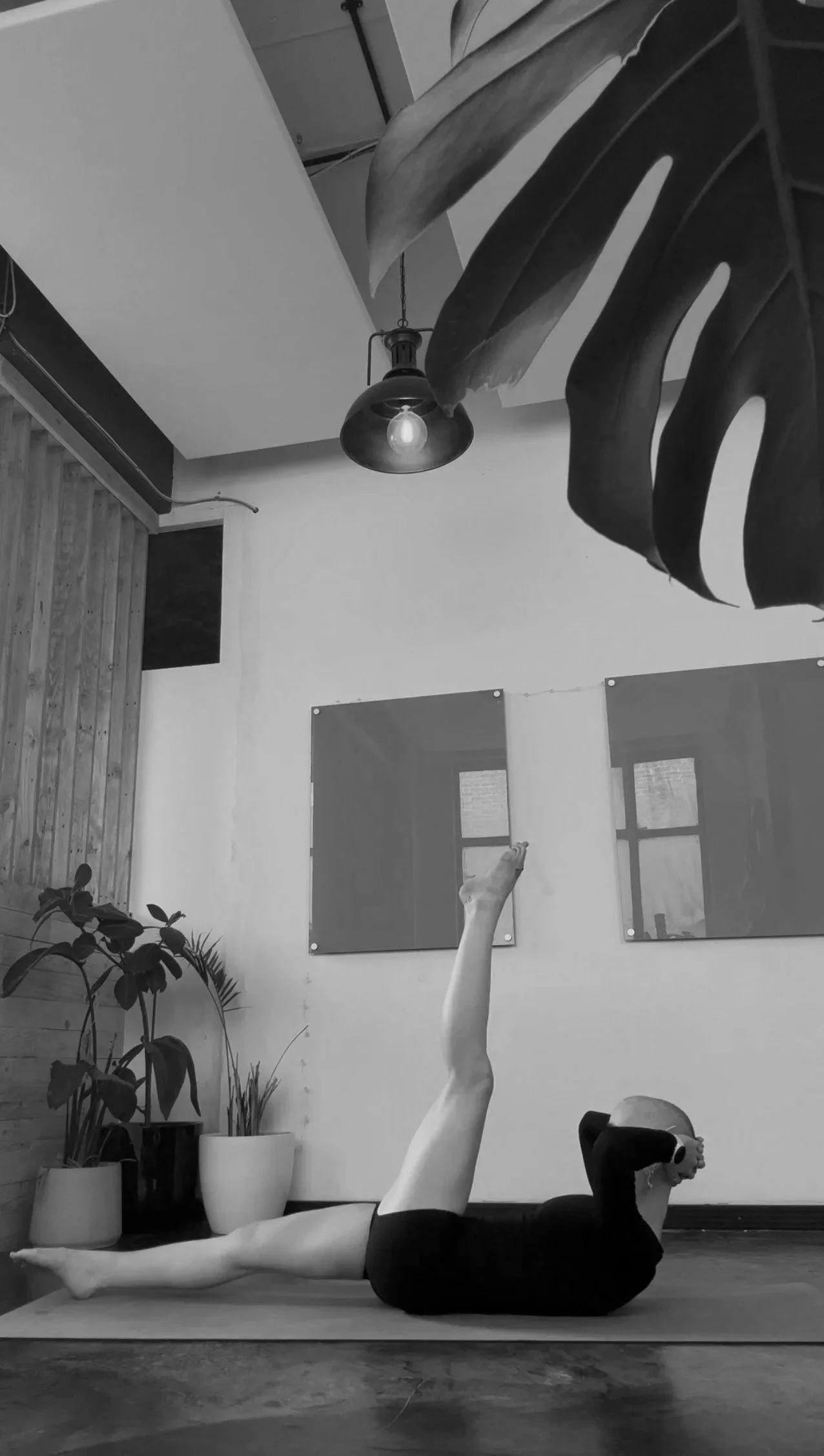 A person practicing yoga in a studio, lying on their side with one leg extended upward, wearing black athletic wear, with potted plants, wall mirrors, and framed artwork in the background.