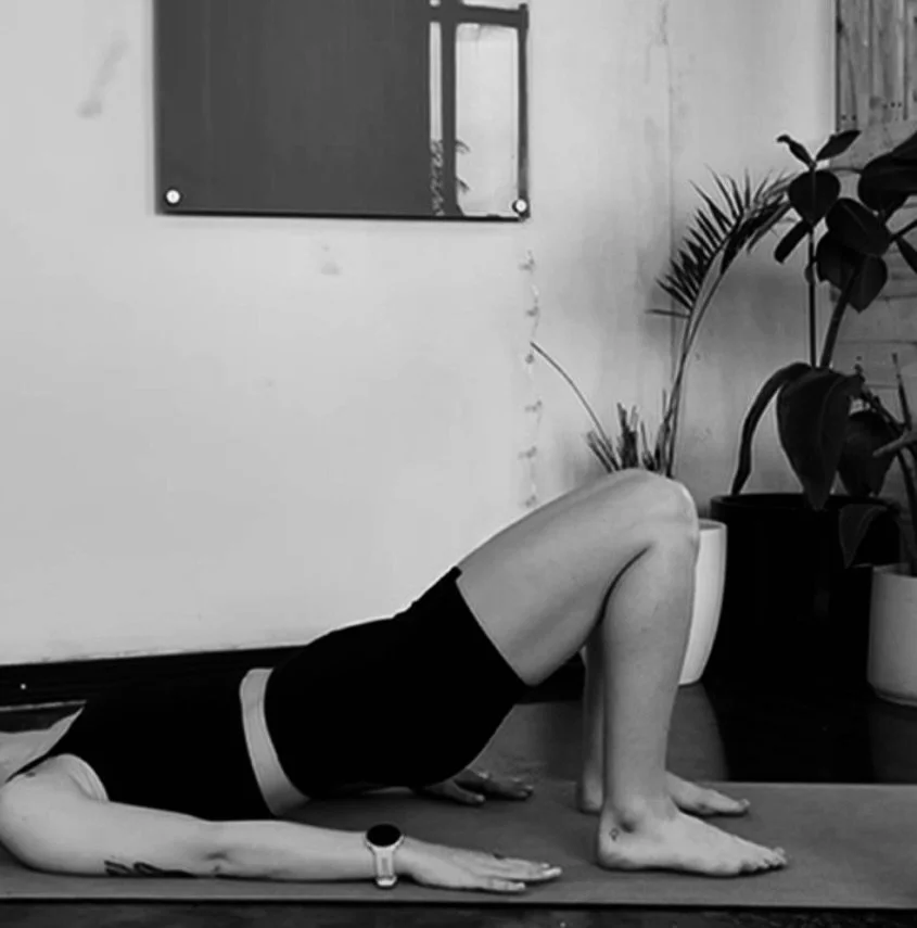 A person lying on a yoga mat in a yoga pose with their knees bent and feet flat, head resting on the floor, wearing a black sports bra and shorts, in a room with potted plants and a wall with a mirror.