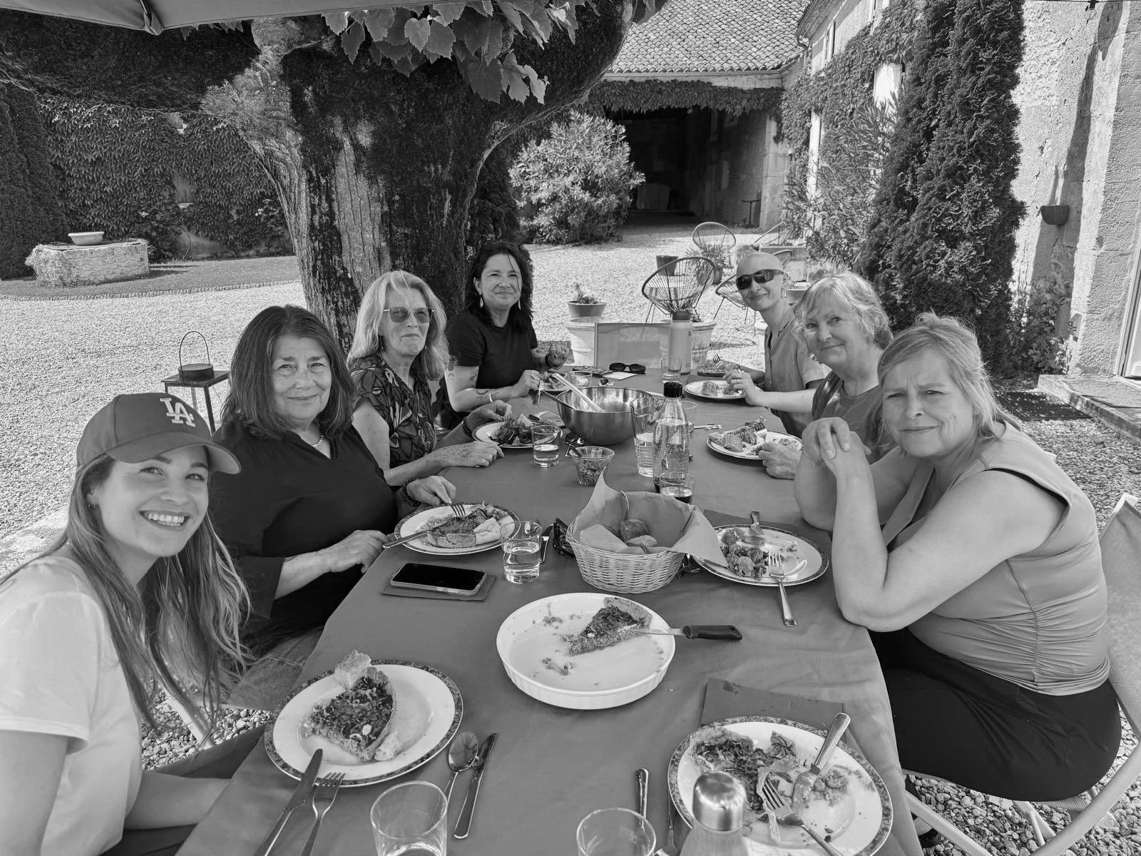A group of eight women sitting around a long outdoor table enjoying a meal, with plates of food, glasses, and a basket of bread, in a courtyard with trees and a building in the background.