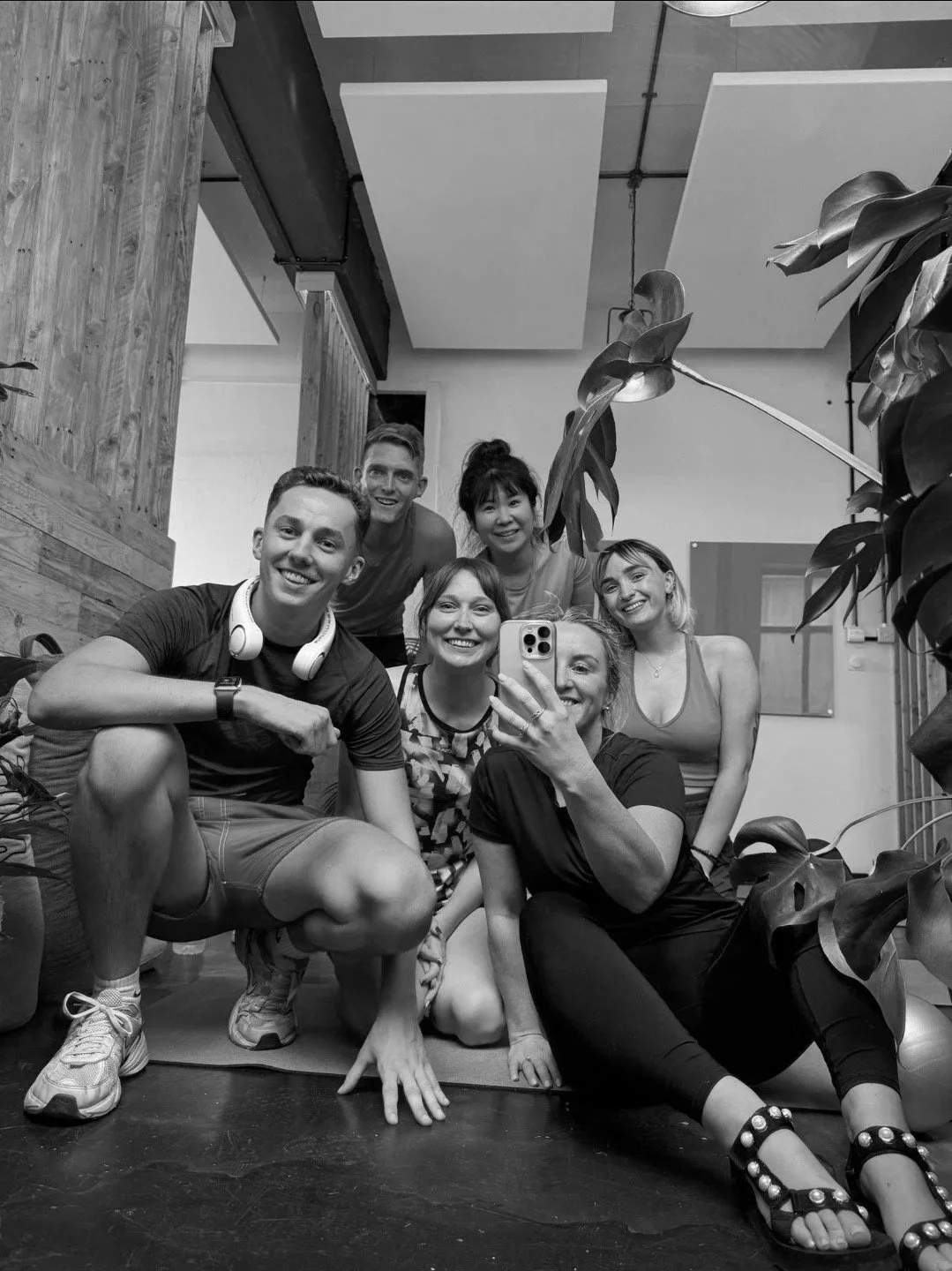 Group of six people taking a selfie in a fitness or yoga studio, some seated and some crouching, with plants and wooden decor in the background.