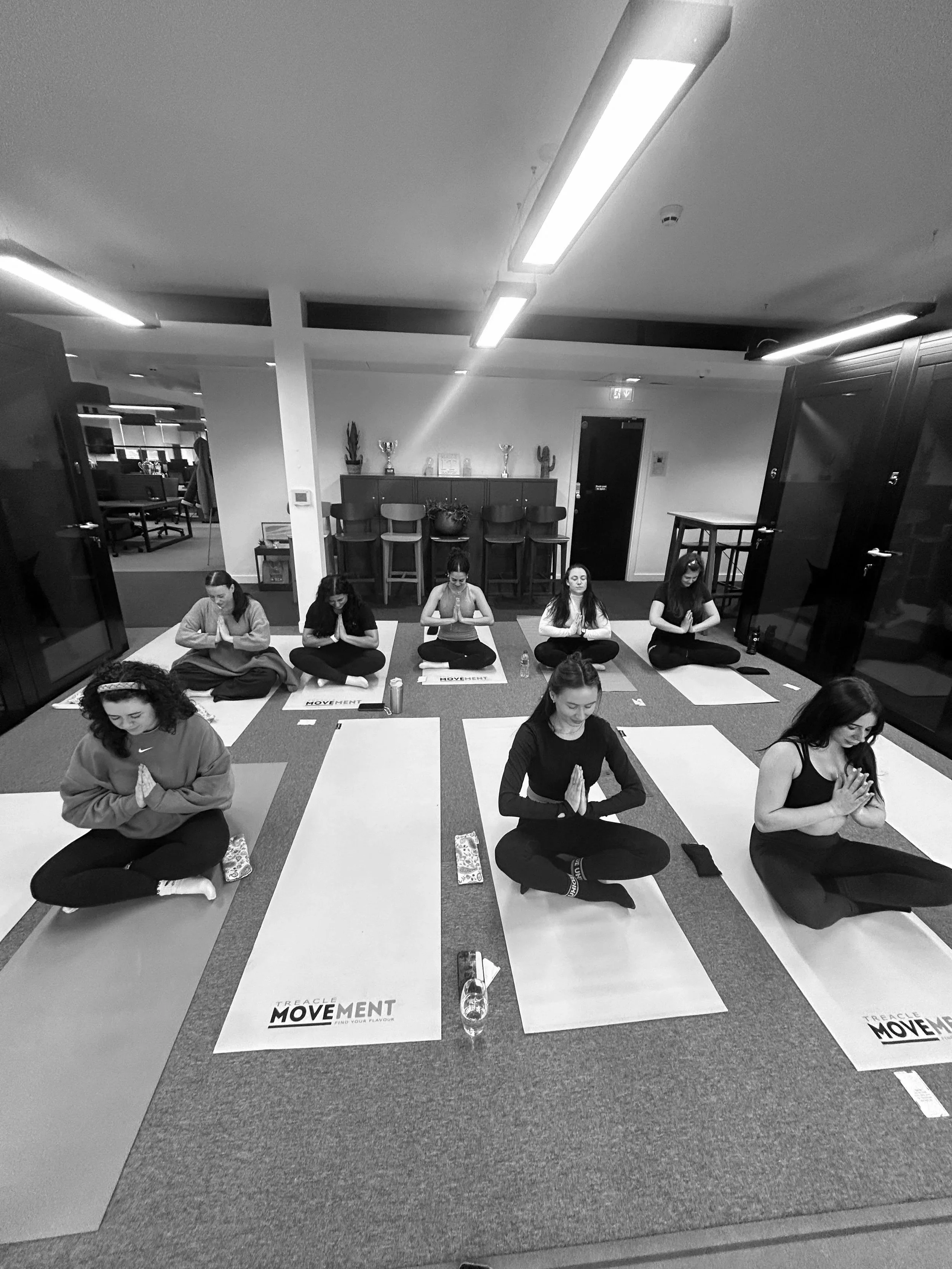 Indoor yoga class with seven women sitting cross-legged on yoga mats, in a meditation pose with hands pressed together in front of their chests. The studio has a carpeted floor, overhead fluorescent lighting, and a wall with decorative items and plan