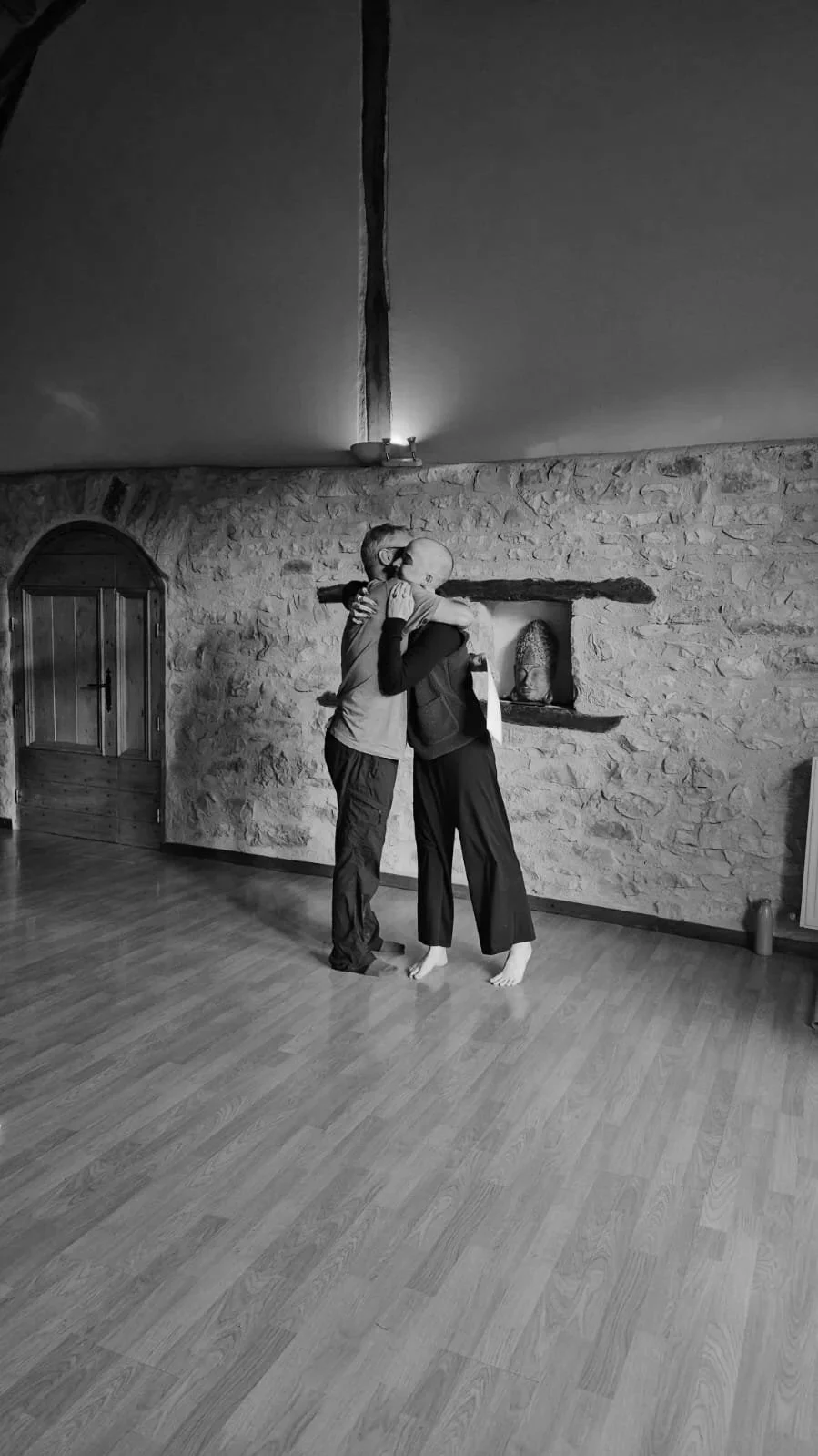 Two people hugging in an indoor room with a stone wall, wooden door, and hardwood floor. The photograph is in black and white.