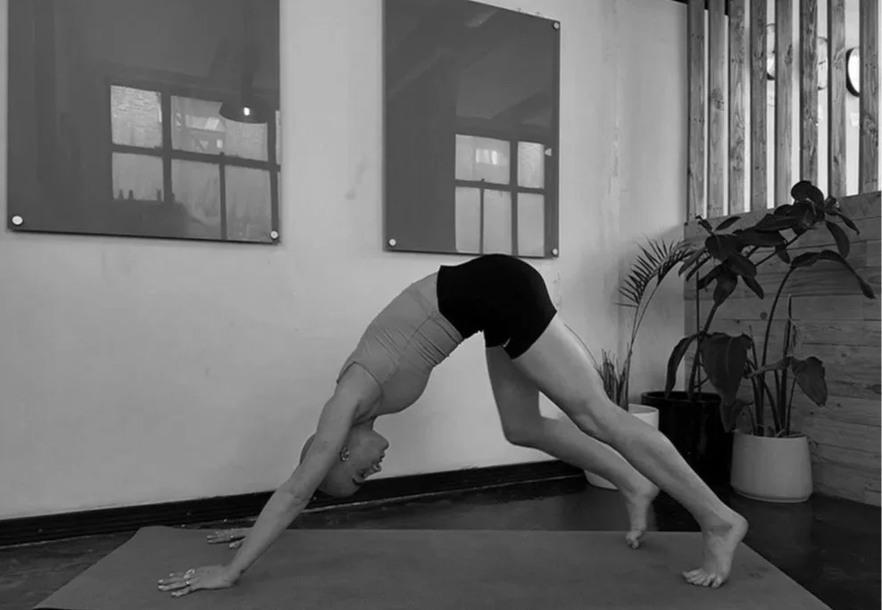 A person practicing yoga in a room, performing a downward-facing dog pose on a yoga mat with potted plants and framed pictures on the wall.