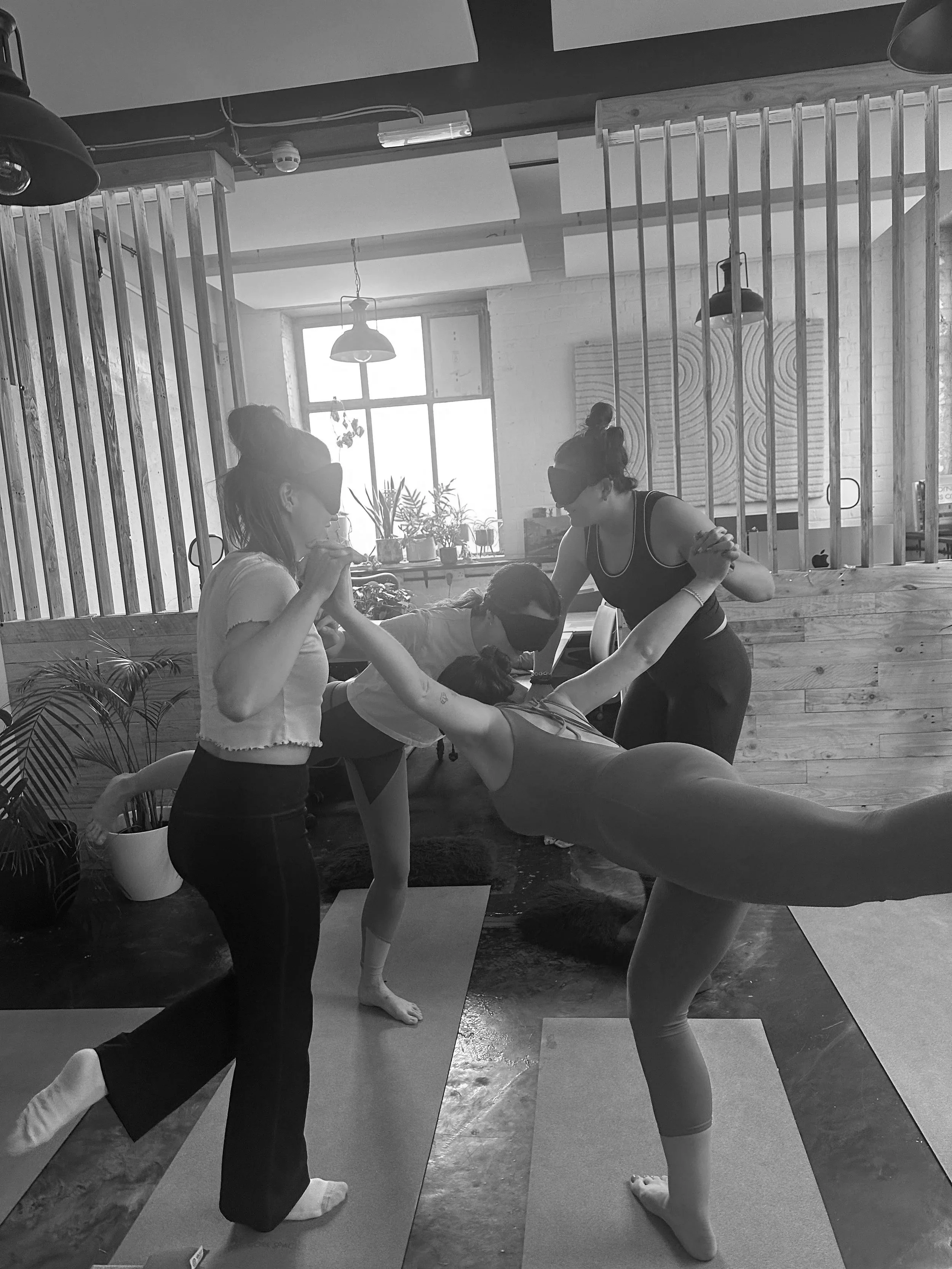 People practicing yoga in a studio, with two women assisting a person in a forward bend pose on mats.