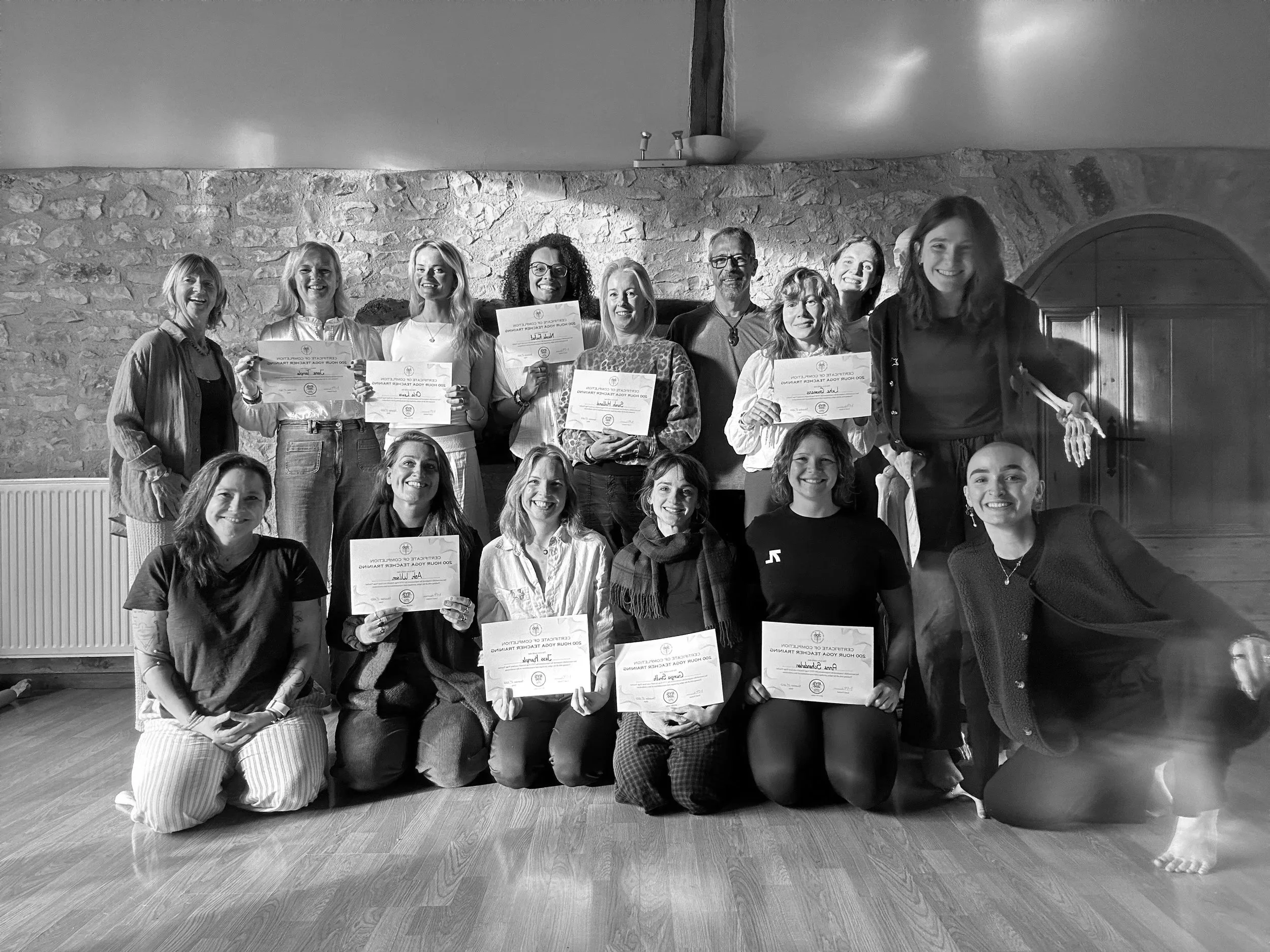 Group of people holding certificates in a room with a stone wall, some women are kneeling in front while others stand behind, all smiling.
