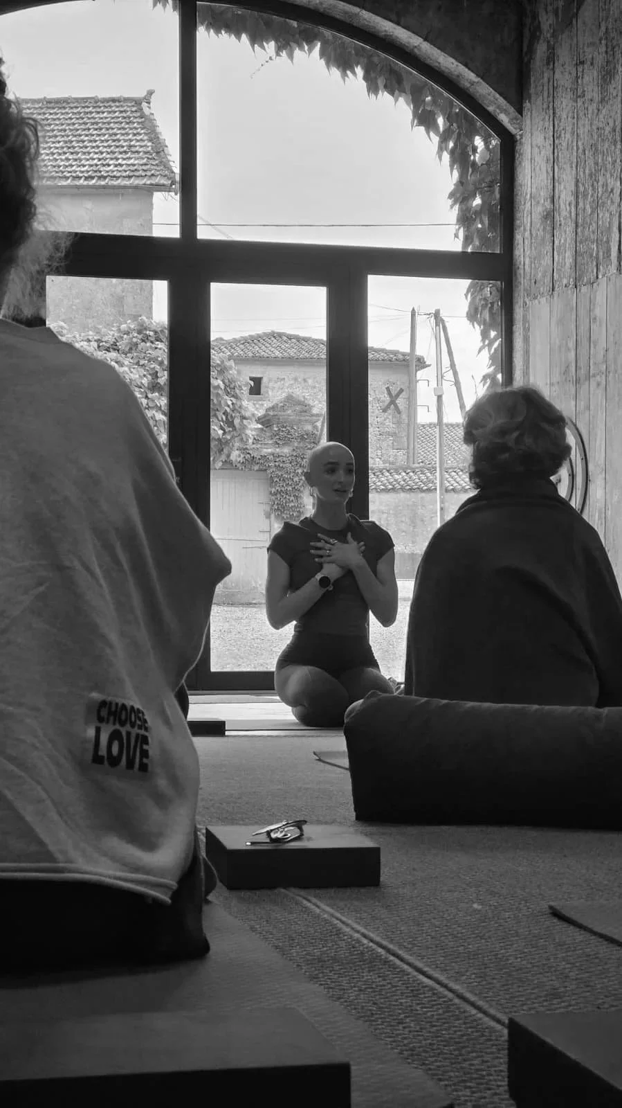 A woman kneeling inside a room with large windows, touching her chest with both hands. Two other women are seated on the floor in front of her, listening to her. The background includes outdoor trees and rooftops.
