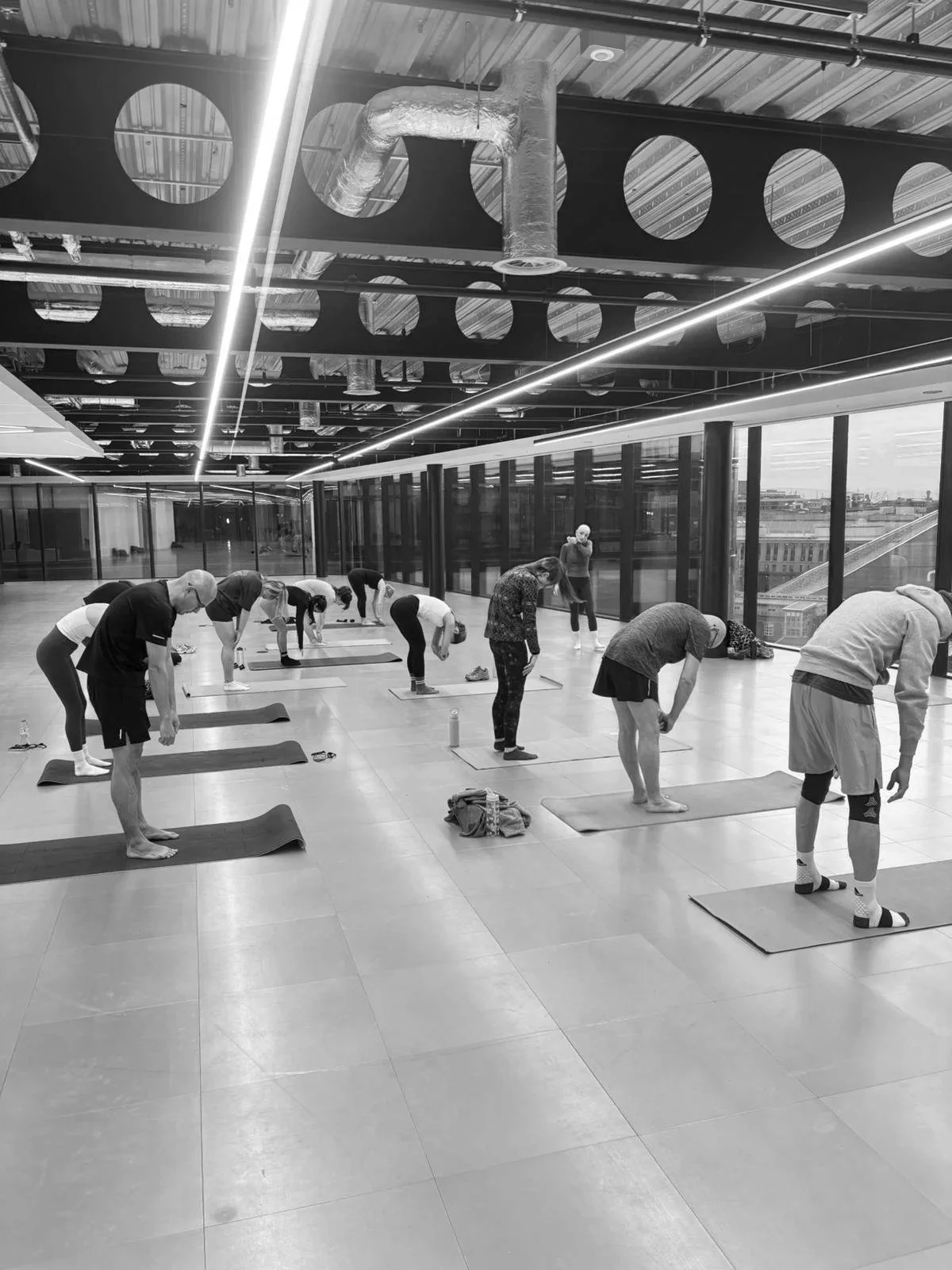 People participating in a yoga class in a spacious, modern indoor space with large windows, exposed ceiling pipes, and an industrial design.