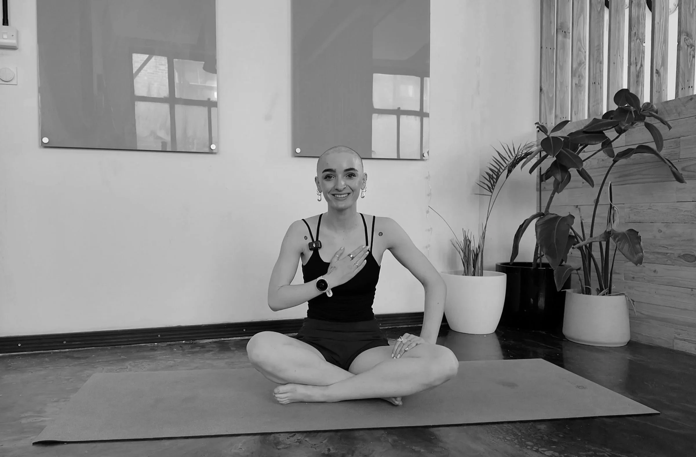 A woman practicing yoga indoors on a yoga mat, sitting cross-legged with one hand on her chest and the other resting on her leg, smiling at the camera, with potted plants and wall art in the background.