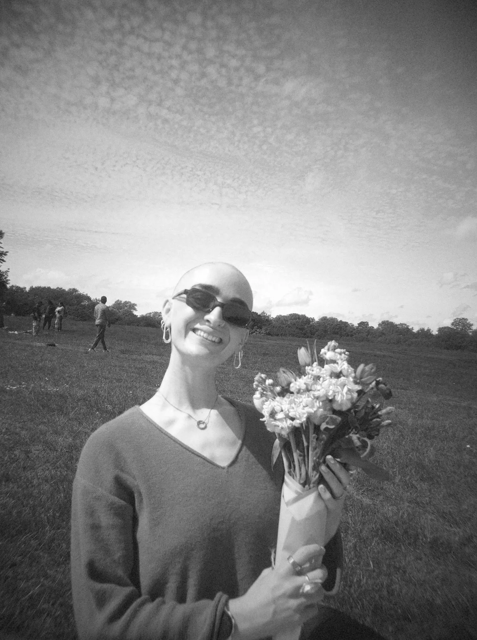A smiling woman with bald head and sunglasses holding a bouquet of flowers outdoors with a group of people in a park and a cloudy sky in the background.