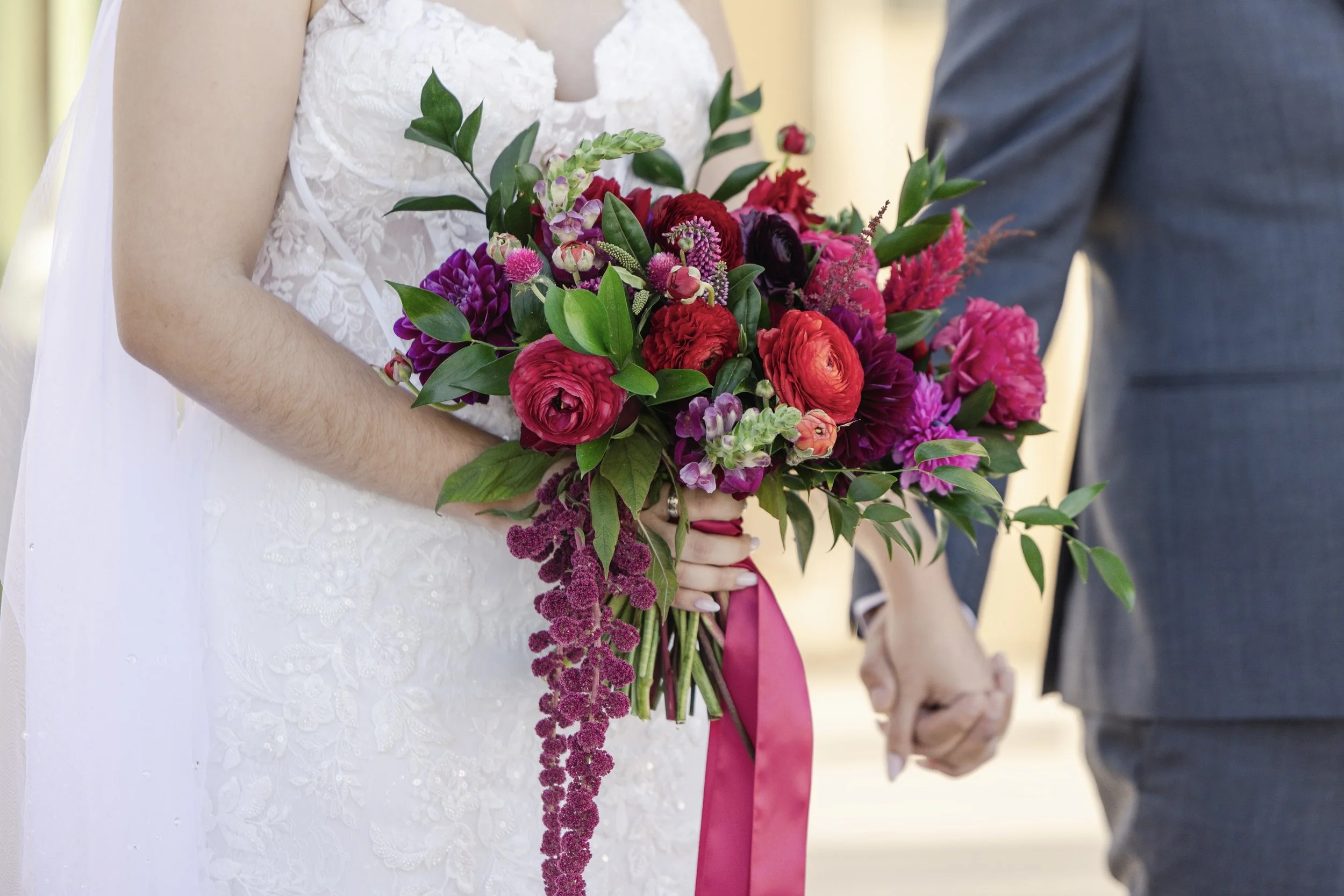 A bride in a white lace dress holding a large bouquet of colorful pink, purple, and red flowers, with greenery, while standing next to a person in a gray suit, both holding hands.