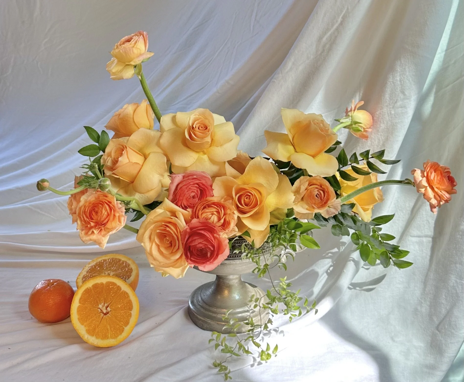 A bouquet of yellow and orange roses in a silver vase, with sliced oranges on a white draped background.