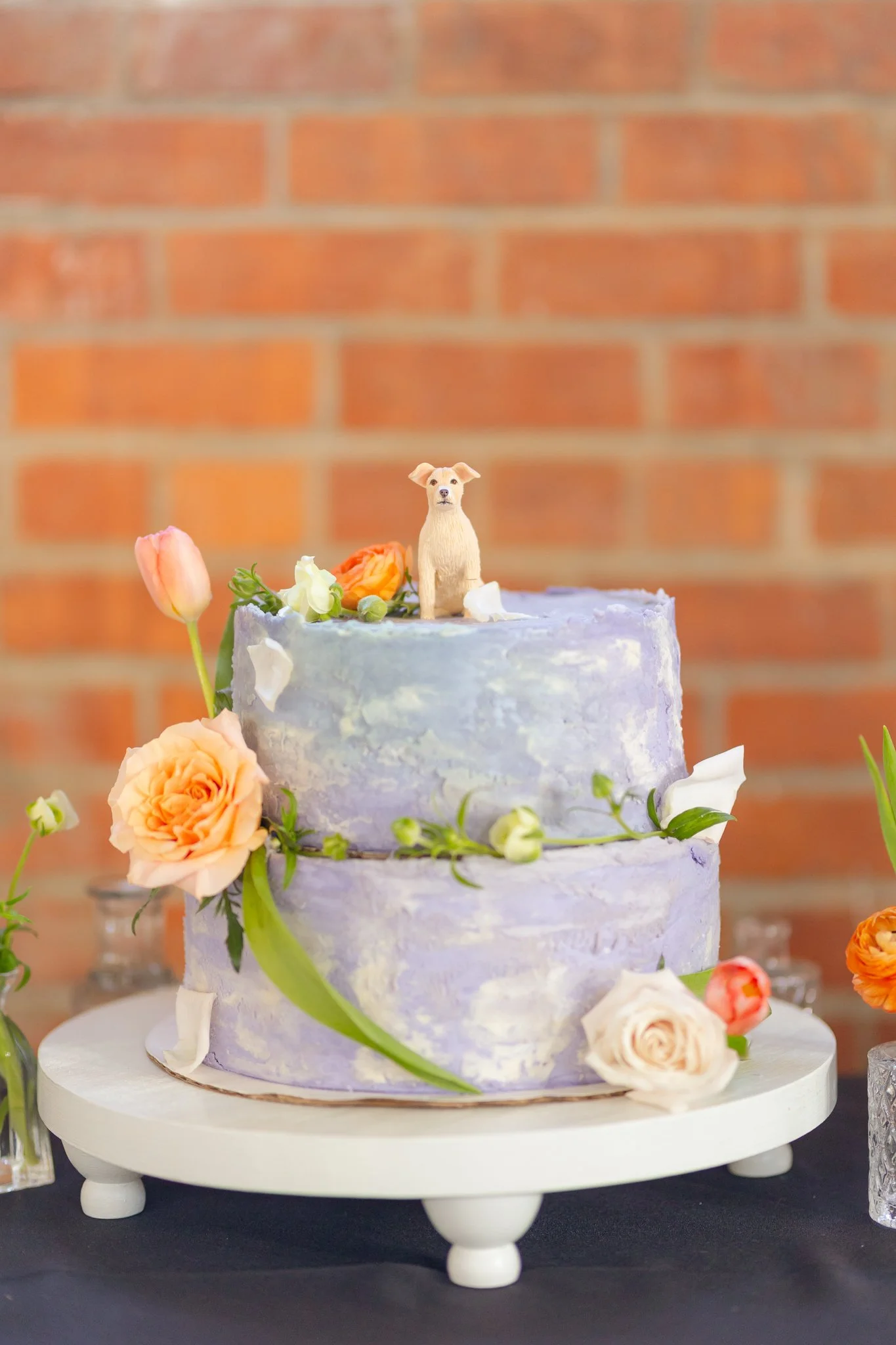 A two-tiered wedding cake decorated with fresh flowers and a small dog figurine on top, set against a brick wall background.
