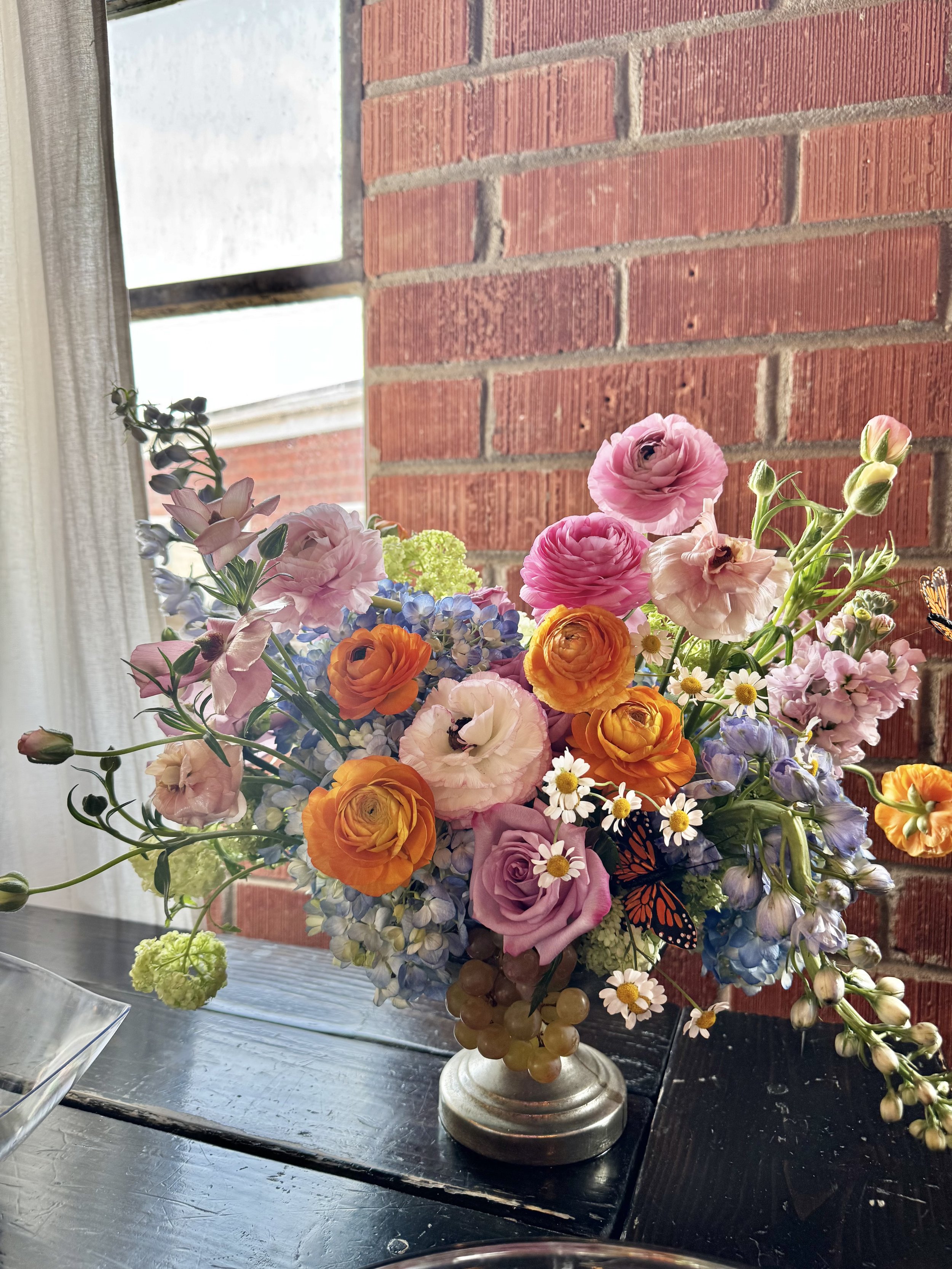 A colorful bouquet of flowers on a table with a brick wall and a window in the background.