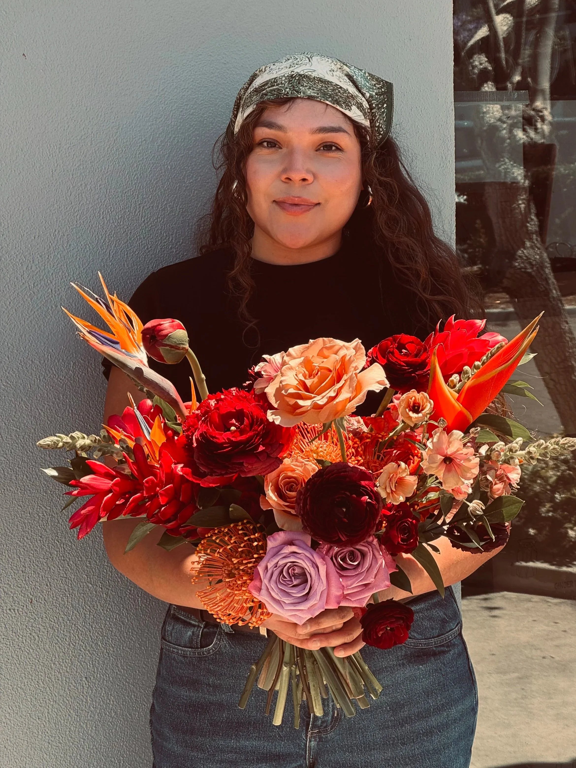 Person in black shirt holding a large colorful bouquet of flowers, wearing a patterned headscarf, standing against a plain wall.