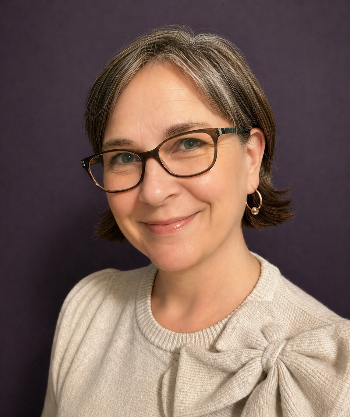 A woman with short brown hair and glasses, smiling, wearing a cream-colored sweater with a bow and a pearl earring, against a dark background.