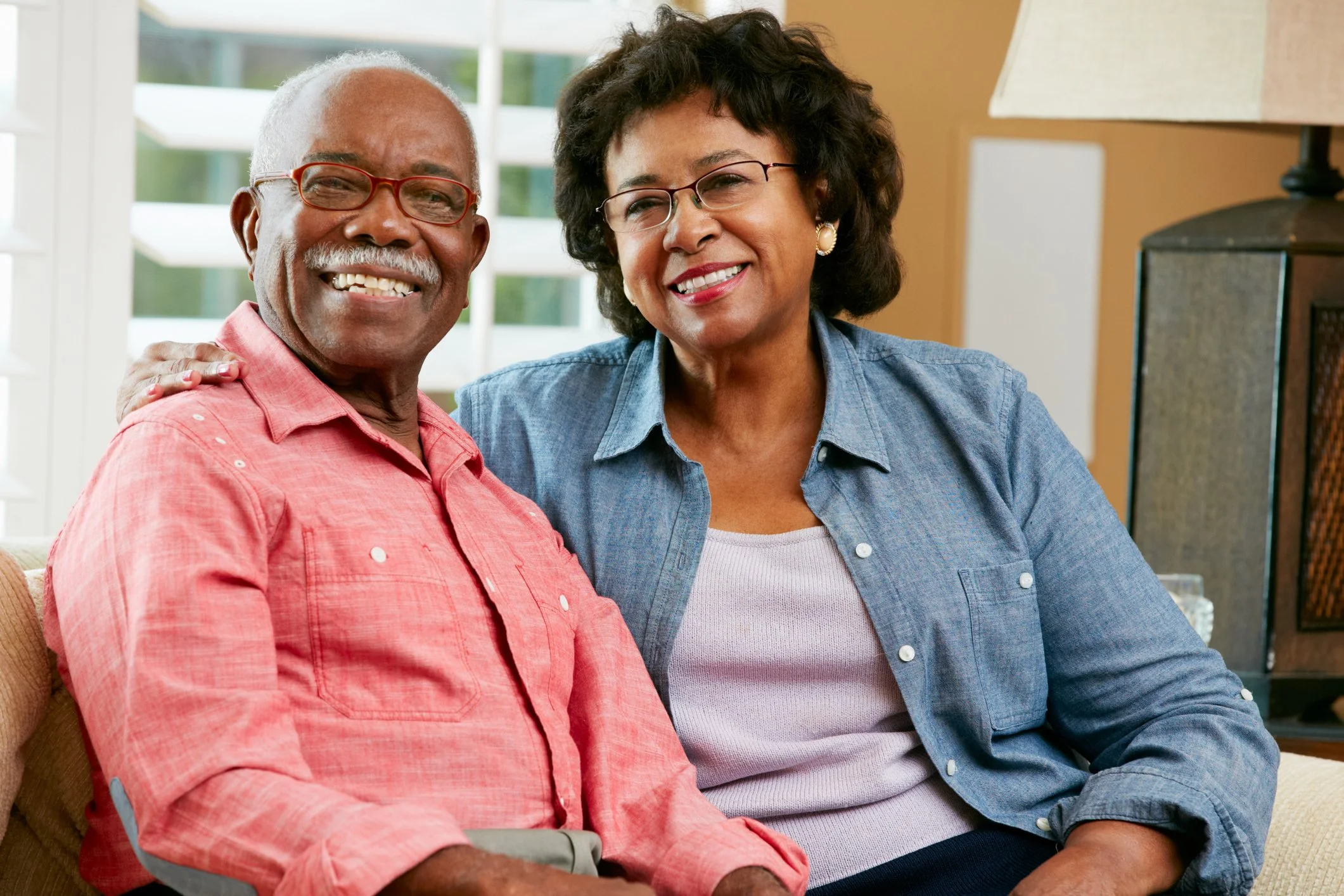 Older man with gray hair and glasses wearing a pink shirt, and an older woman with glasses and curly hair wearing a gray shirt, sitting close together on a beige couch, smiling in a well-lit living room.