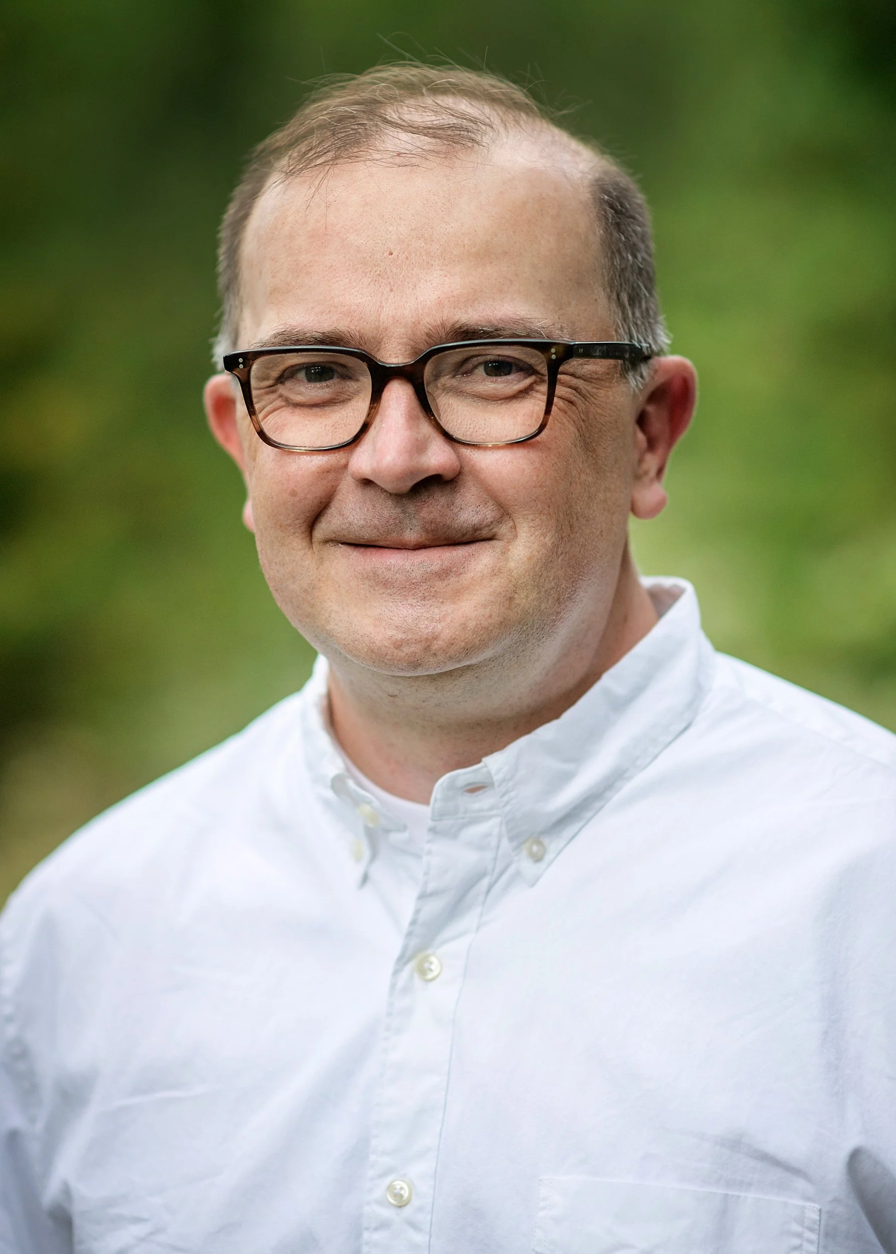 Close-up of a man with glasses, smiling, wearing a white collared shirt, outdoors with blurred green background.