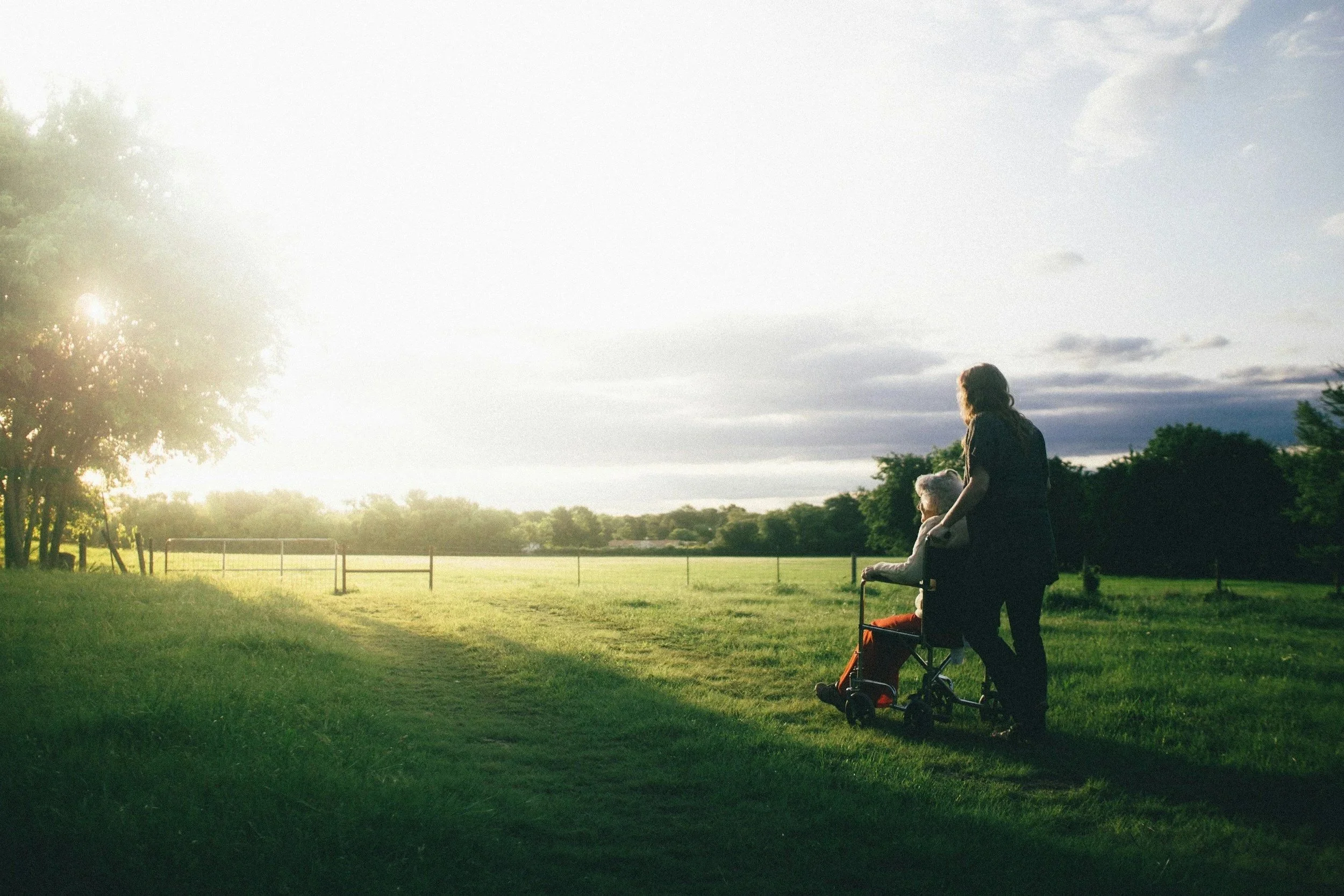 A woman is pushing a young girl in a wheelchair across a grassy field at sunset. There are trees and a fence in the background, and the sky is partly cloudy.