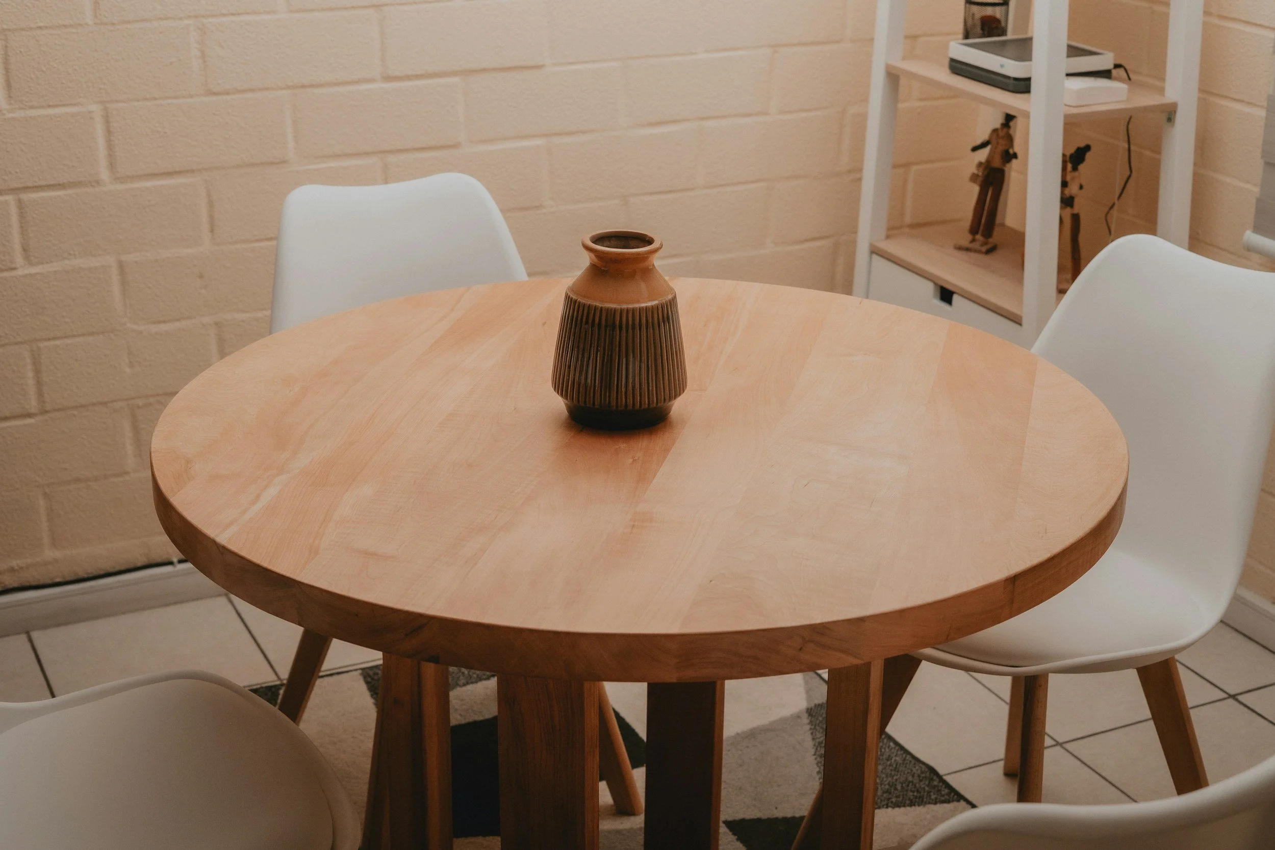 A round wooden dining table with a brown striped ceramic vase in the center, surrounded by three white chairs, in a room with a cream brick wall and a beige rug.