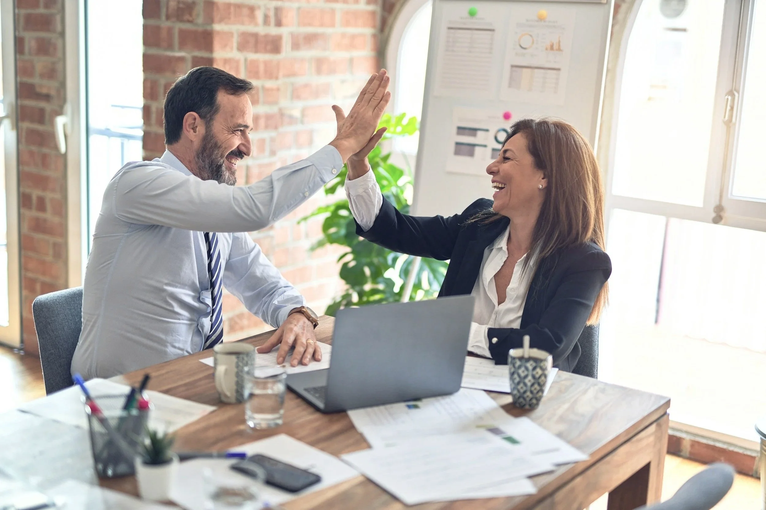 Two colleagues, a man and a woman, smiling and giving each other a high five in an office, with papers, a laptop, and coffee cups on the desk.