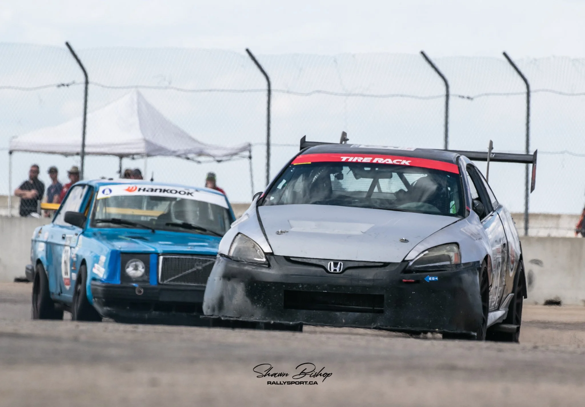 Close-up of a black Honda race car with a large rear wing, driven during a racing event, with a blue race car visible in the background and people standing behind a fence.