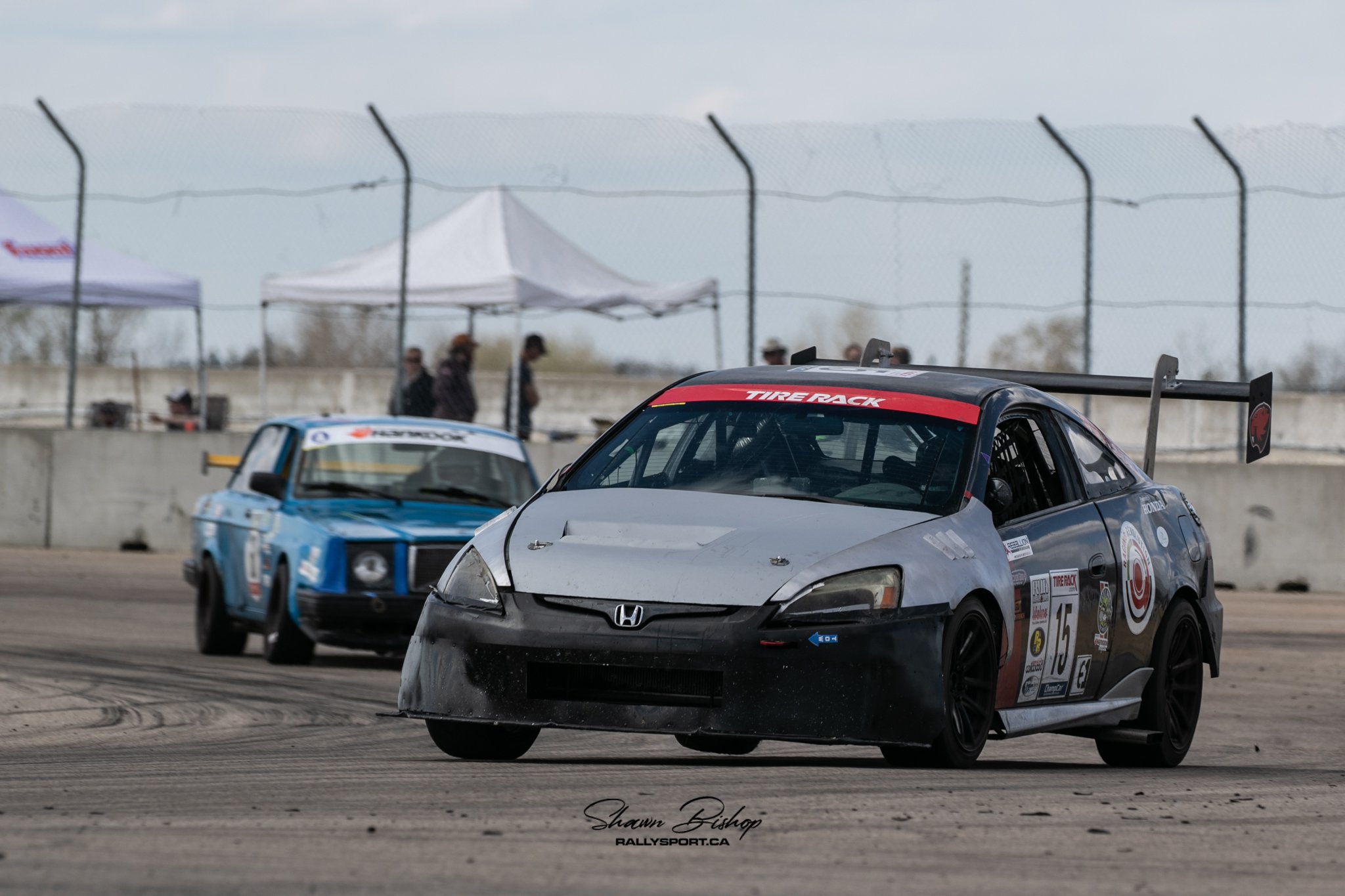 Close-up of a black Honda race car with a large rear spoiler, number 15 on the side, on a race track. In the background, there are other race cars, people, tents, and a chain-link fence under cloudy skies.