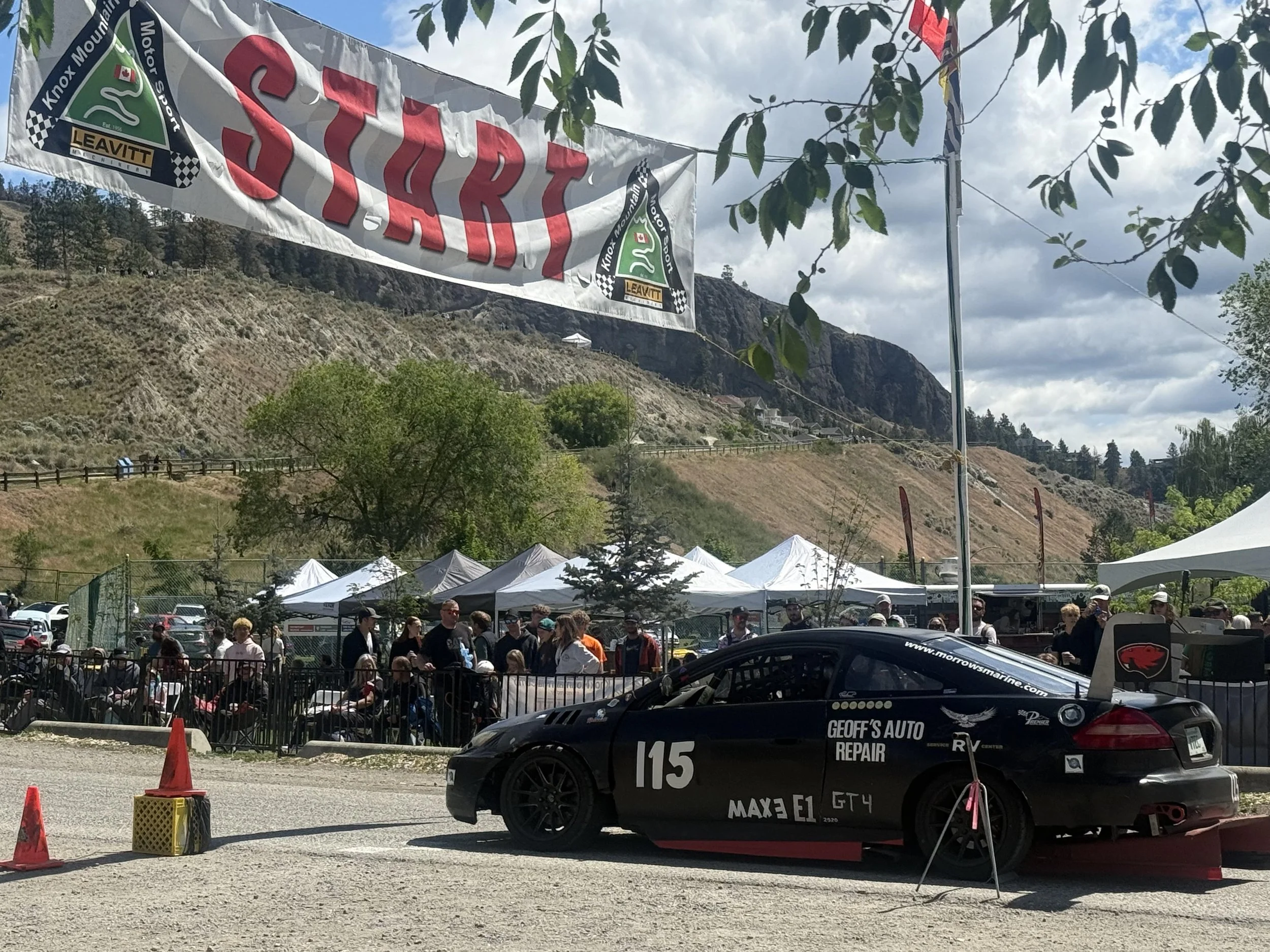 A propped race car with racing decals parked in front of a crowd at an outdoor event, with tents and mountains in the background.