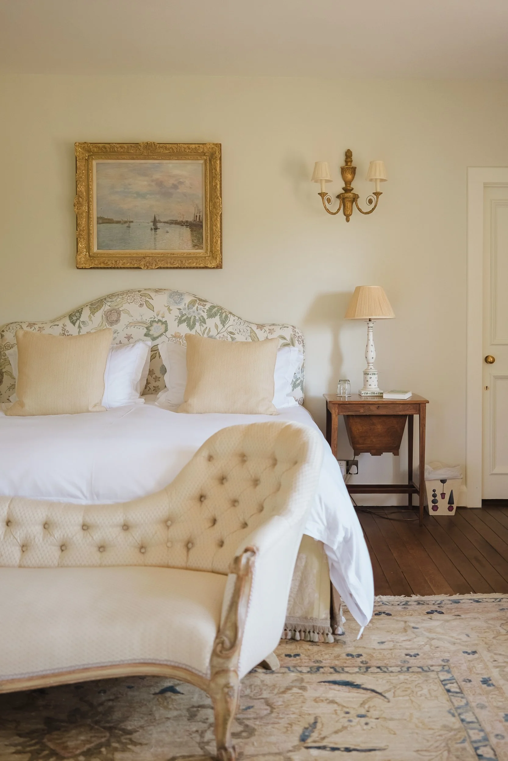 A bedroom with a vintage cream-colored tufted chaise lounge at the foot of a bed with floral-patterned headboard, three pillows, and white bedding. A wooden nightstand holds a beige lamp and a glass of water. Artwork of a harbor scene hangs above the bed, and a wall sconce with two lamps is mounted on the wall.